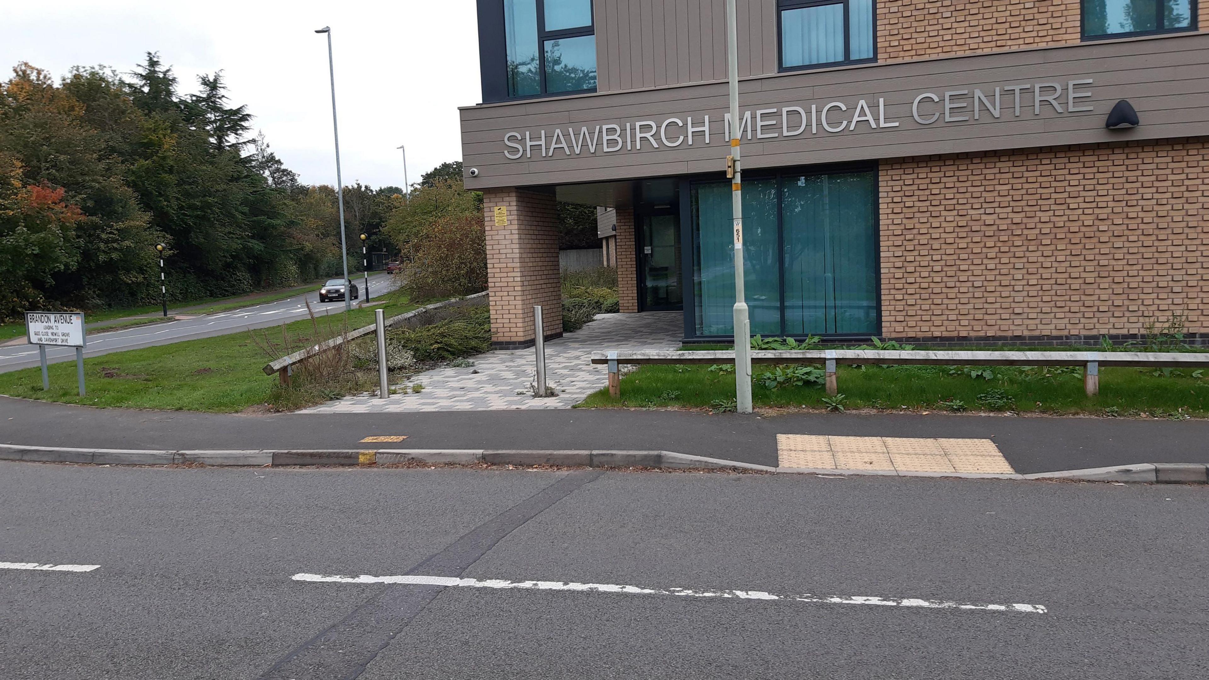 A modern brick building with a brown band of cladding around its middle on which "Shawbirch Medical Centre" is spelled out in silver metallic lettering. The medical centre is on the junction of two roads, with a pedestrian crossing visible on one.  