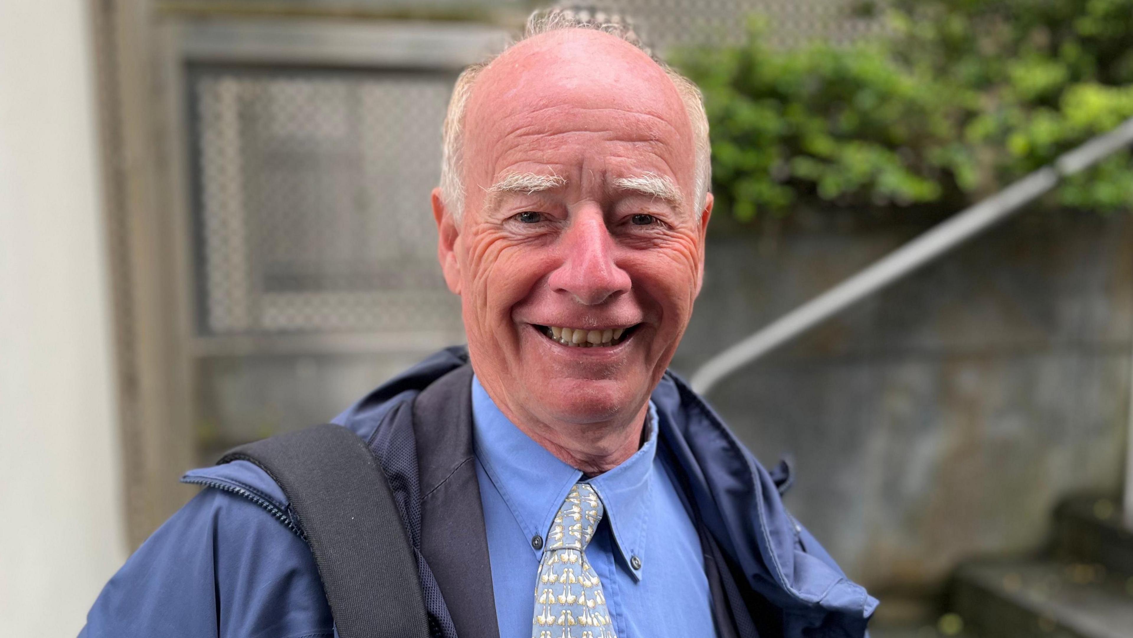 Deputy George Oswald looking towards the camera. He's smiling and has a bald head with white hair on the side of his face.
He is wearing a blue overcoat, with a blue shirt and a blue and white tie.