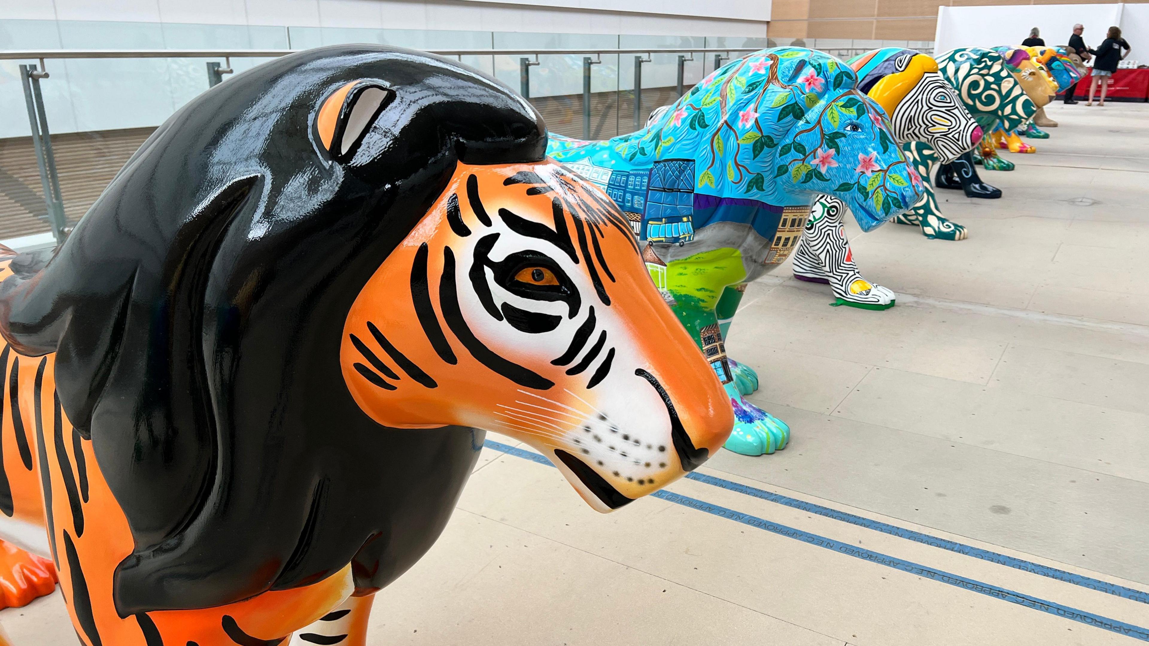 A row of the brightly coloured lions inside in a large warehouse. The one closest to the camera is covered with a tiger print, while the others are all multicoloured.