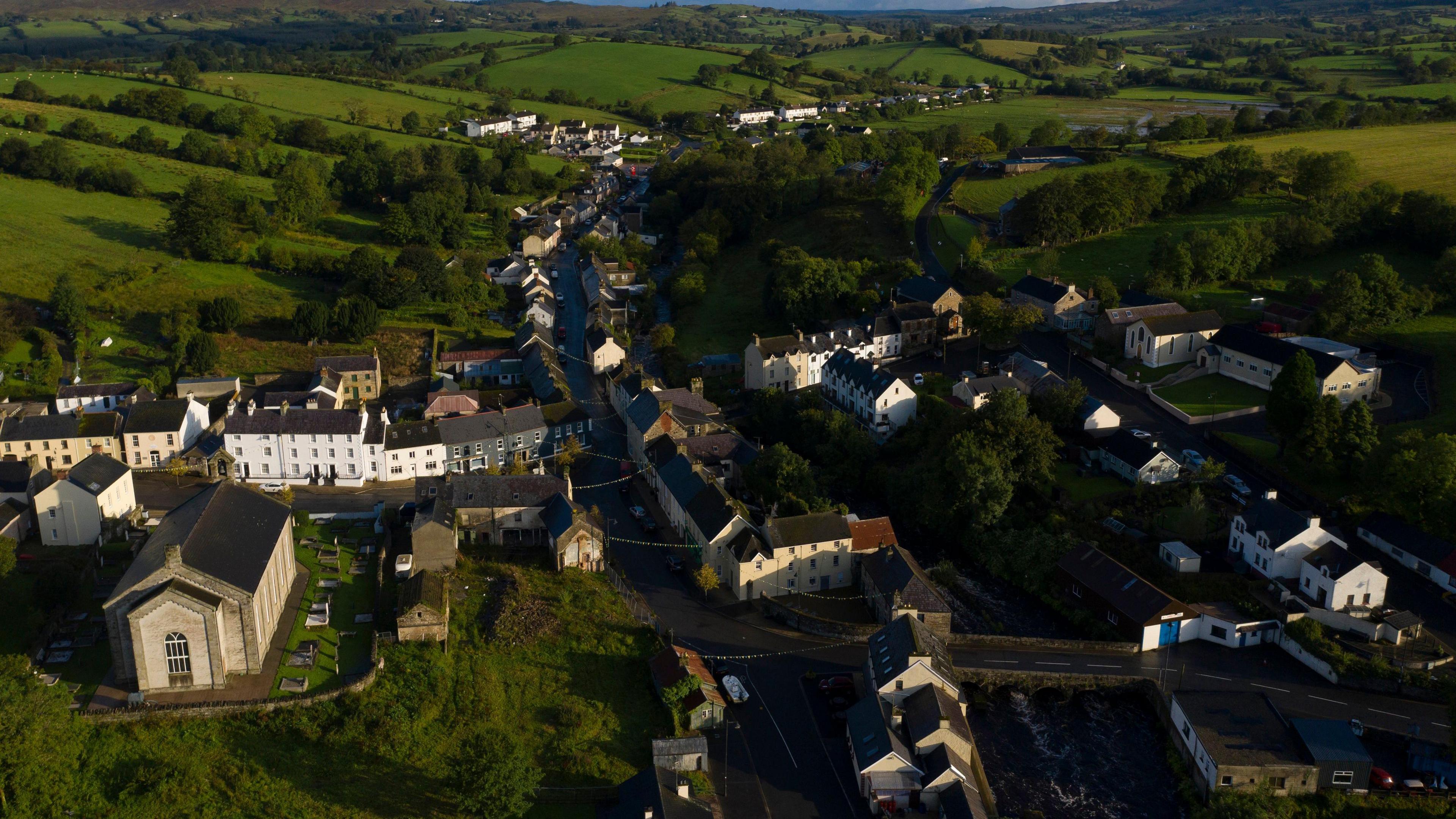 An aerial view of the village of Pettigo, surrounded by green fields.  There is a large church in the right side of the photo which is in County Donegal. An arched stone bridge in the left of the photo marks the gateway into County Fermanagh. 