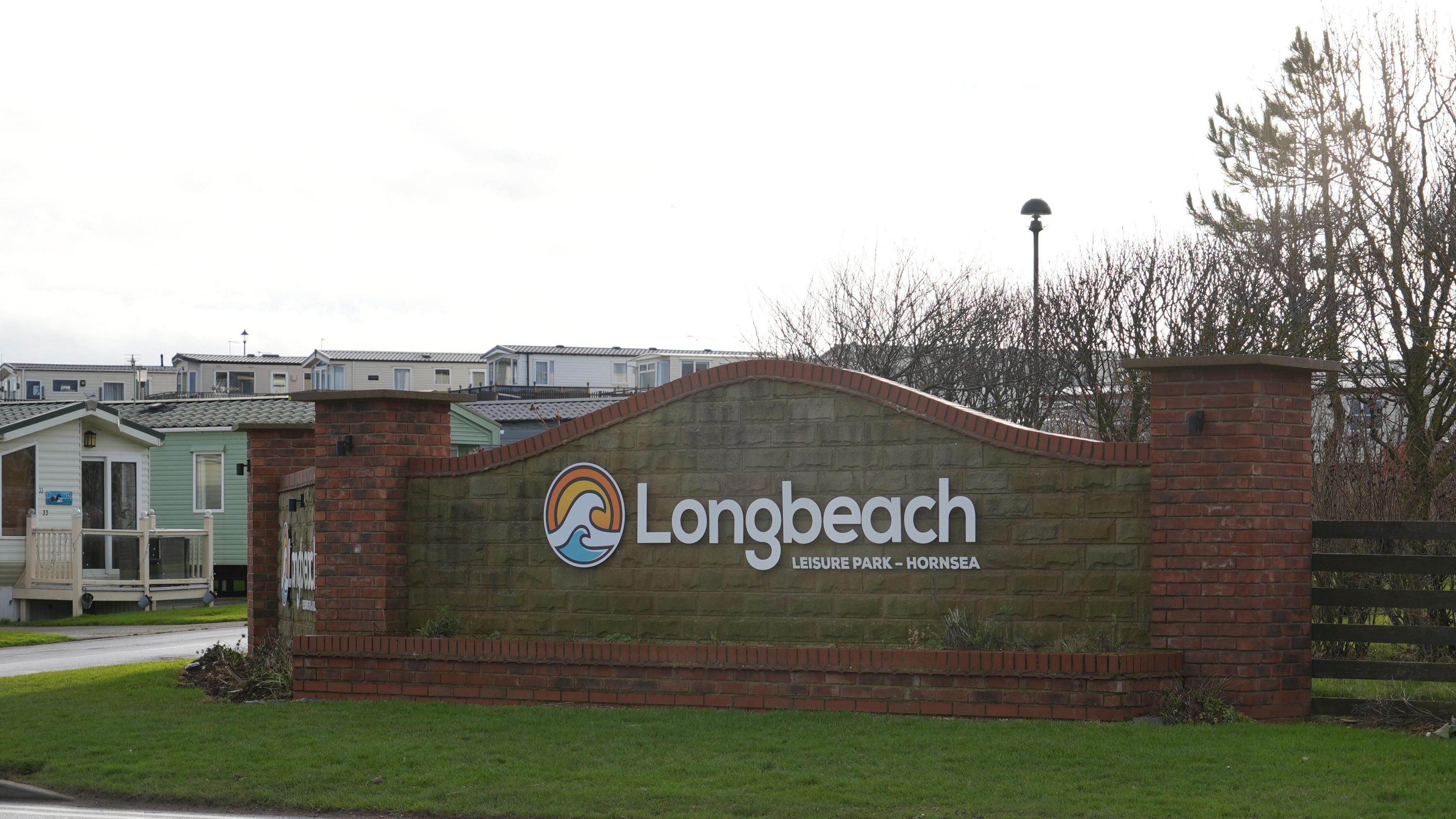 The entrance to the Longbeach Leisure Park. A brick and stone wall stands on a grass verge with the name of the holiday park in large white letters. Several caravans are visible behind the wall.