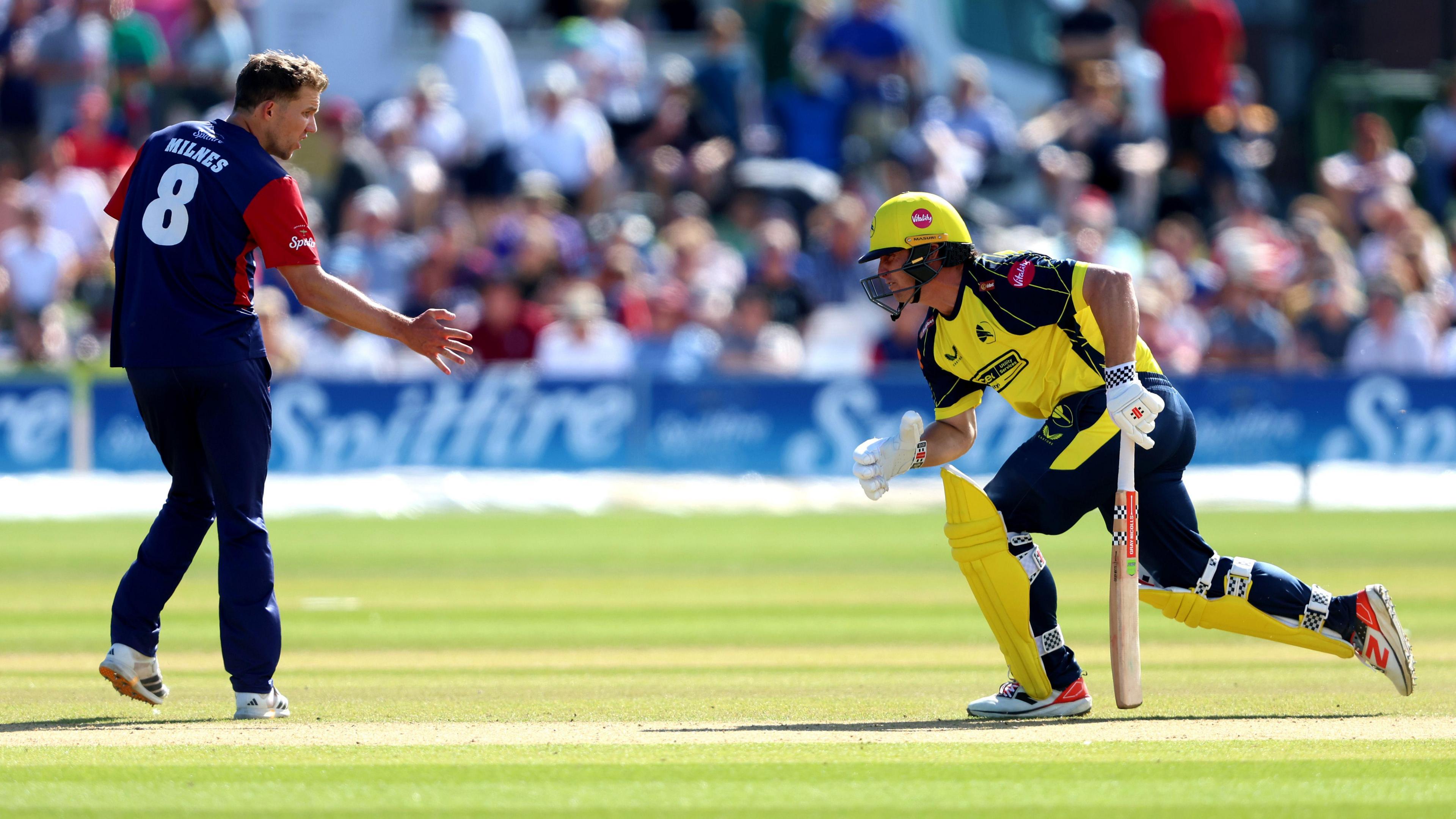 Hampshire Hawks batter Hilton Cartwright running across a wicket during a T20 Blast match against Kent Spitfires while wearing a predominantly yellow kit and helmet with dark blue, approaching an opposition player wearing a blue and red kit