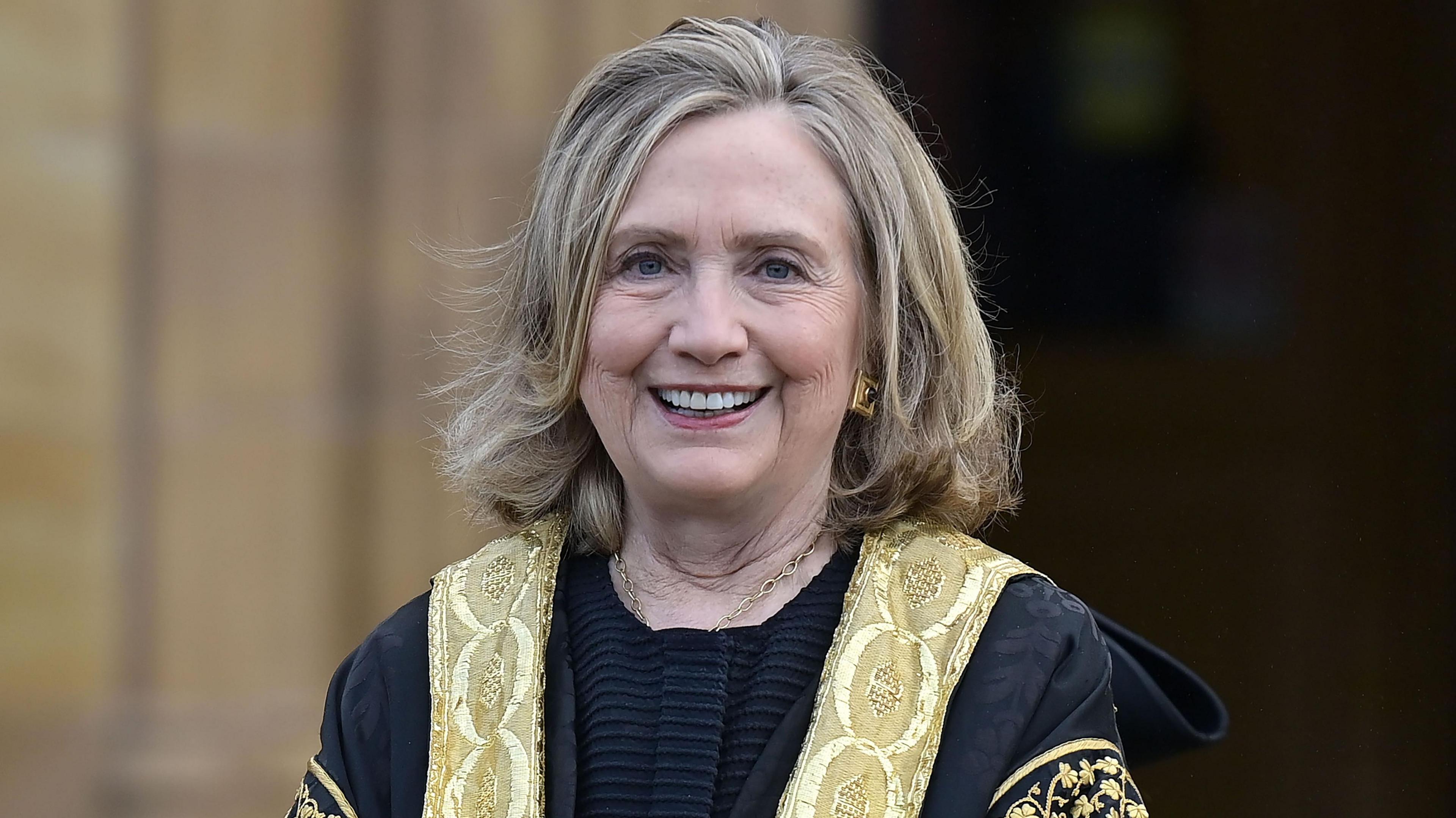 Hillary Clinton wearing a gold and black outfit. She is smiling at the camera. She has blonde hair.