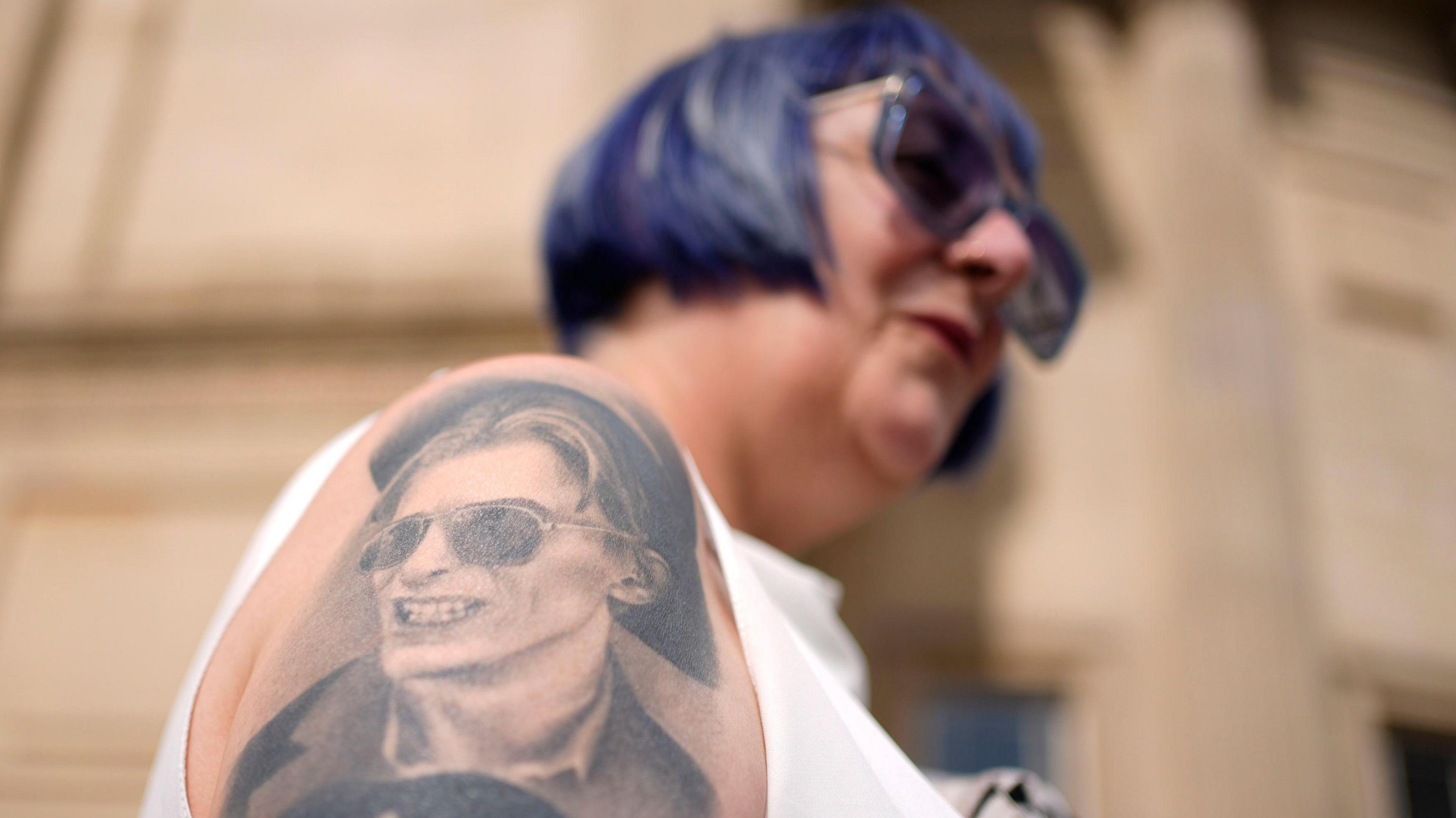 A woman with purple hair in a bob style and purple sunglasses, shows her David Bowie tattoo, an image of the singer wearing a hat and sunglasses, on her arm outside the David Bowie World Fan Convention