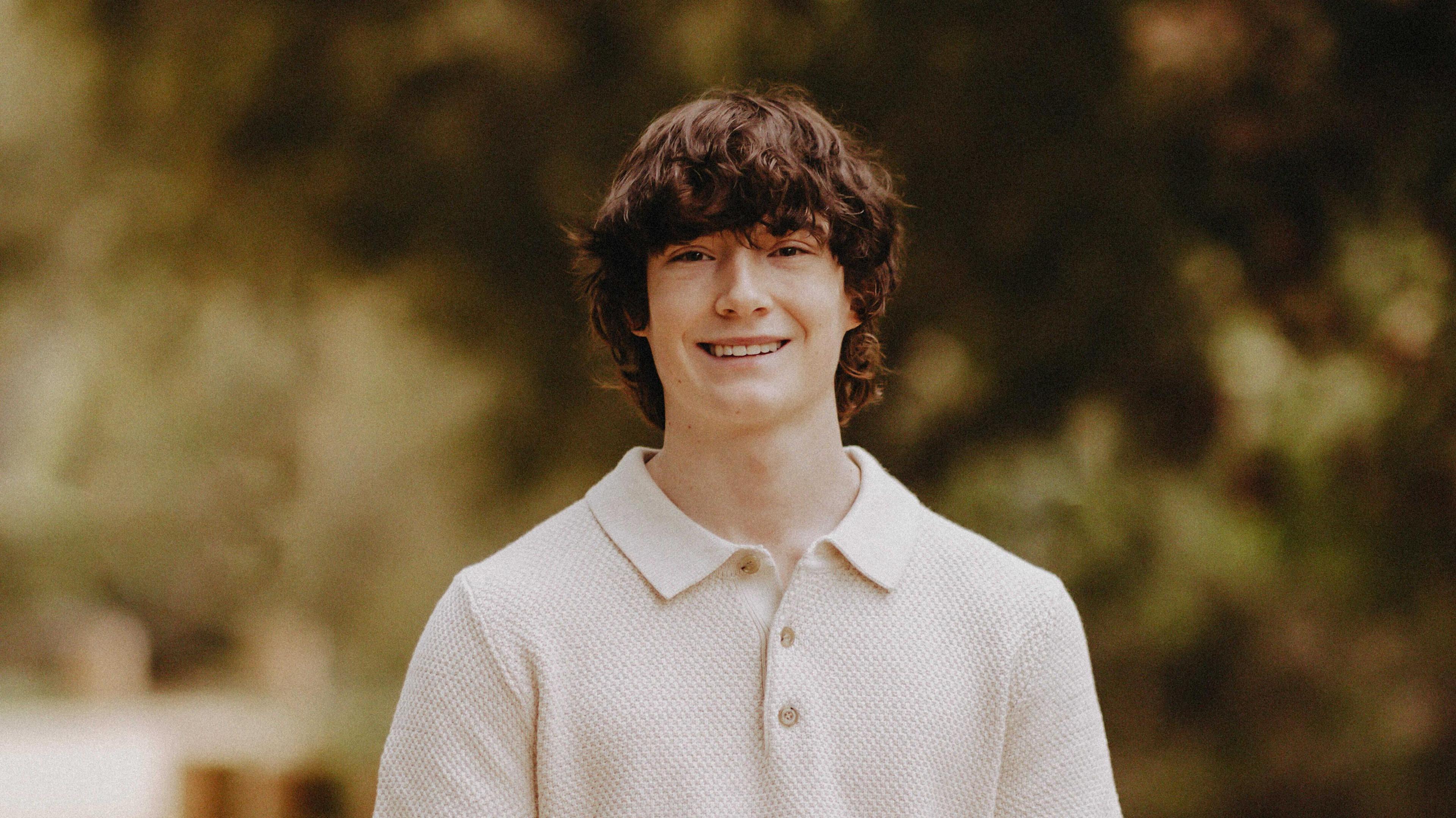 A photo of Adam Raine. He has long, brown shaggy hair that is wavy. He is seen smiling, wearing a knit collared shirt with three buttons at the top. Behind him is a blurred background of foliage.