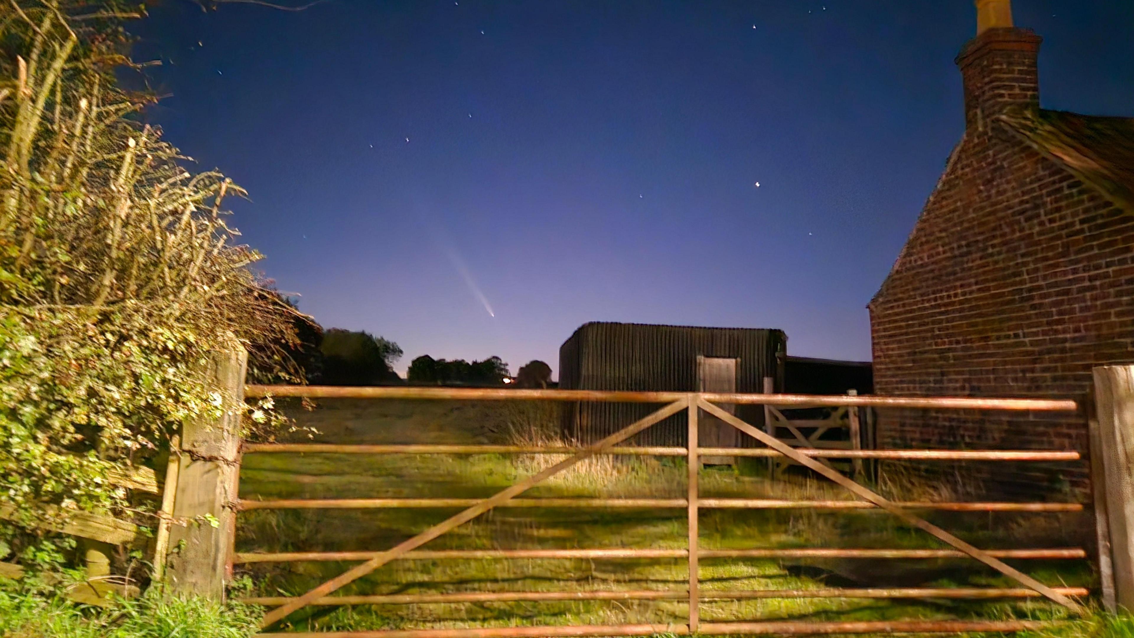 The comet shooting through a inky blue sky, with a farm gate immediately in the foreground.