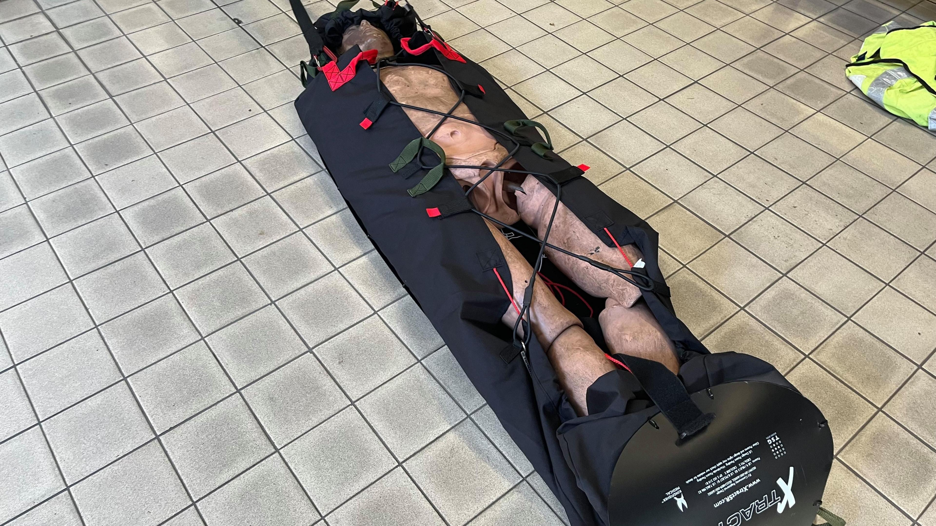 A burned dummy in a black stretcher bag on a tiled floor.