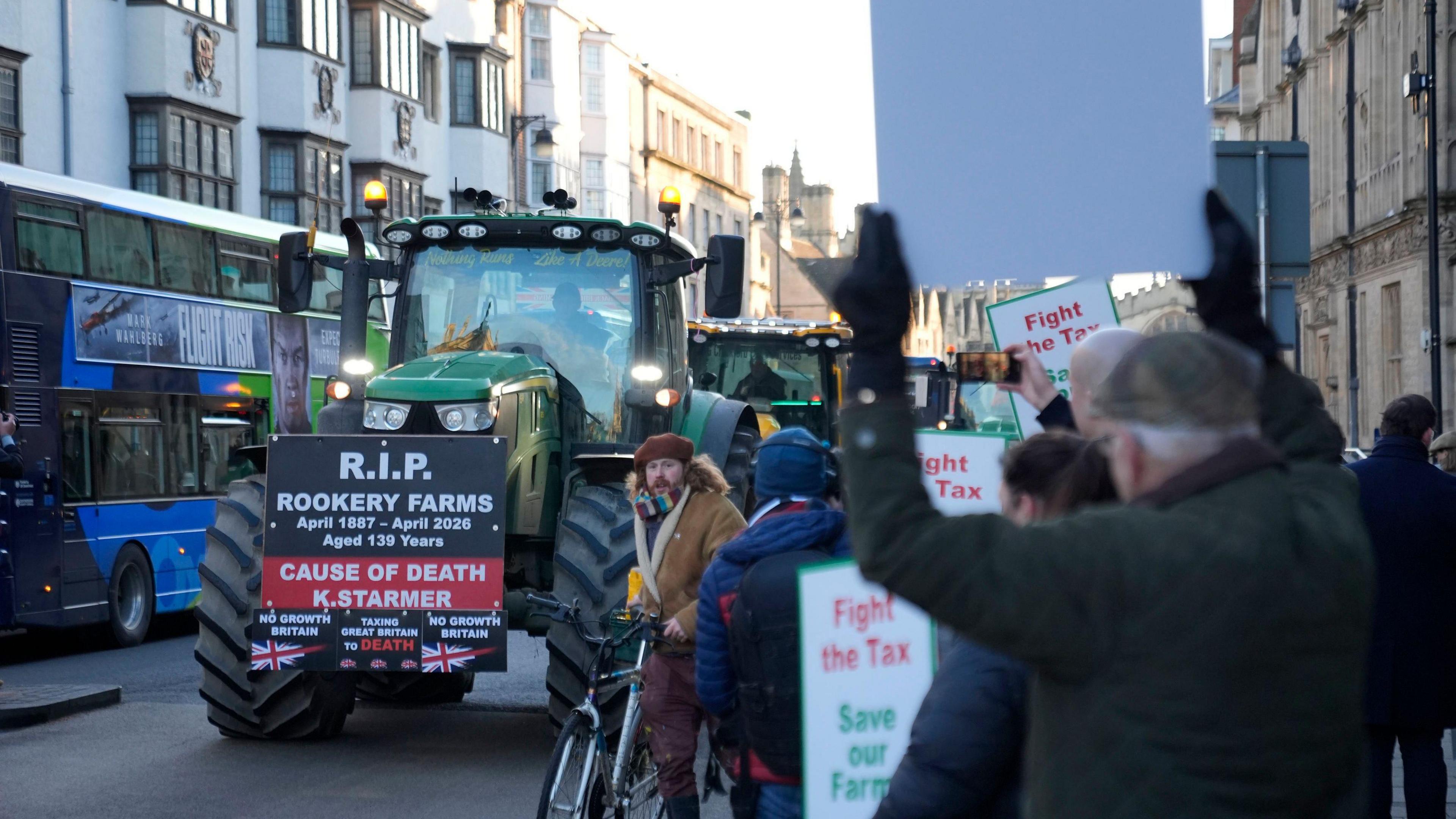 A large green tractor with a large sign on the front of it is driving past people holding placards.