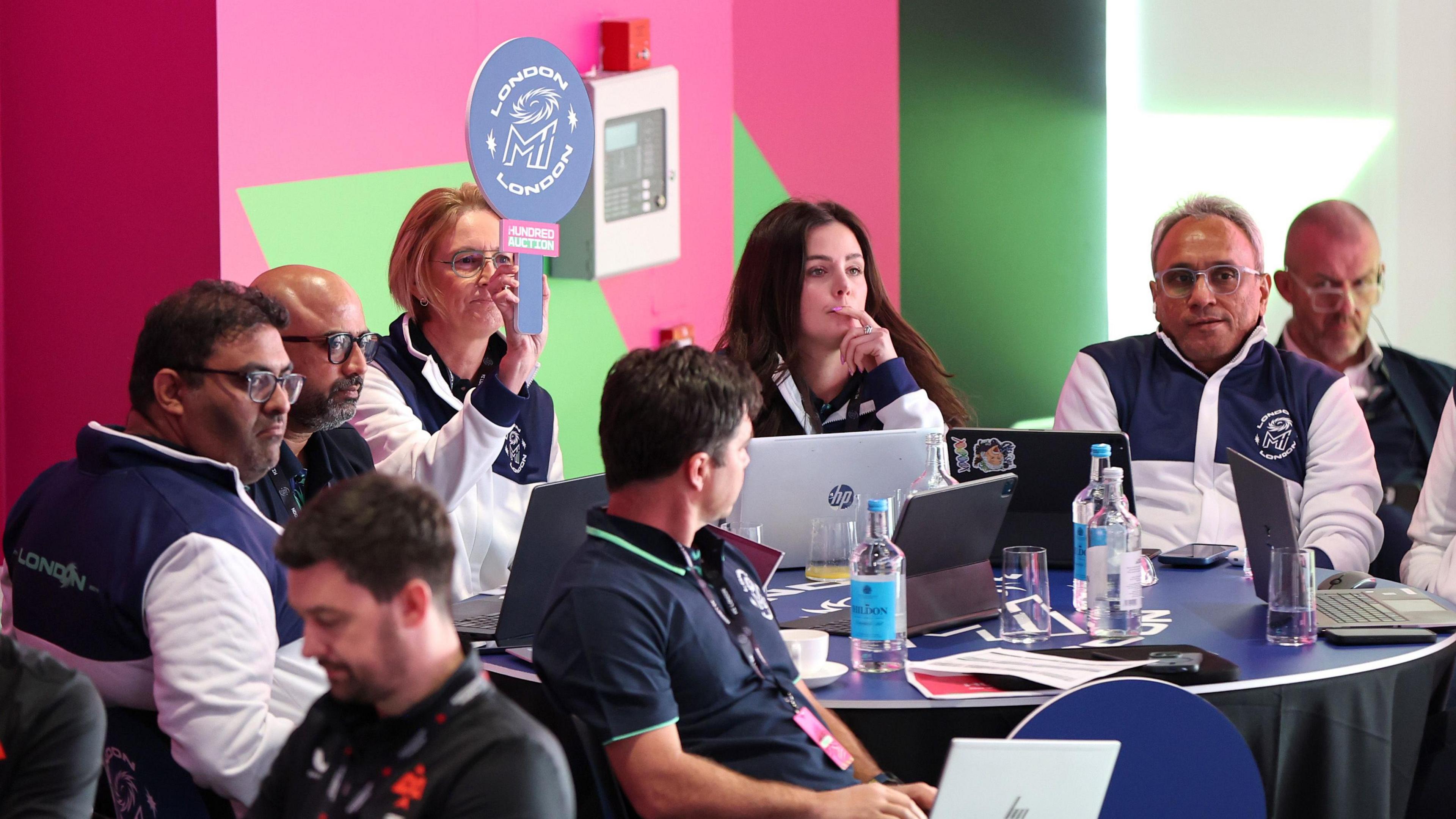 MI London management group sit around a table, meanwhile head coach Lisa Keightley holds up a paddle to signify a bid for a player. 