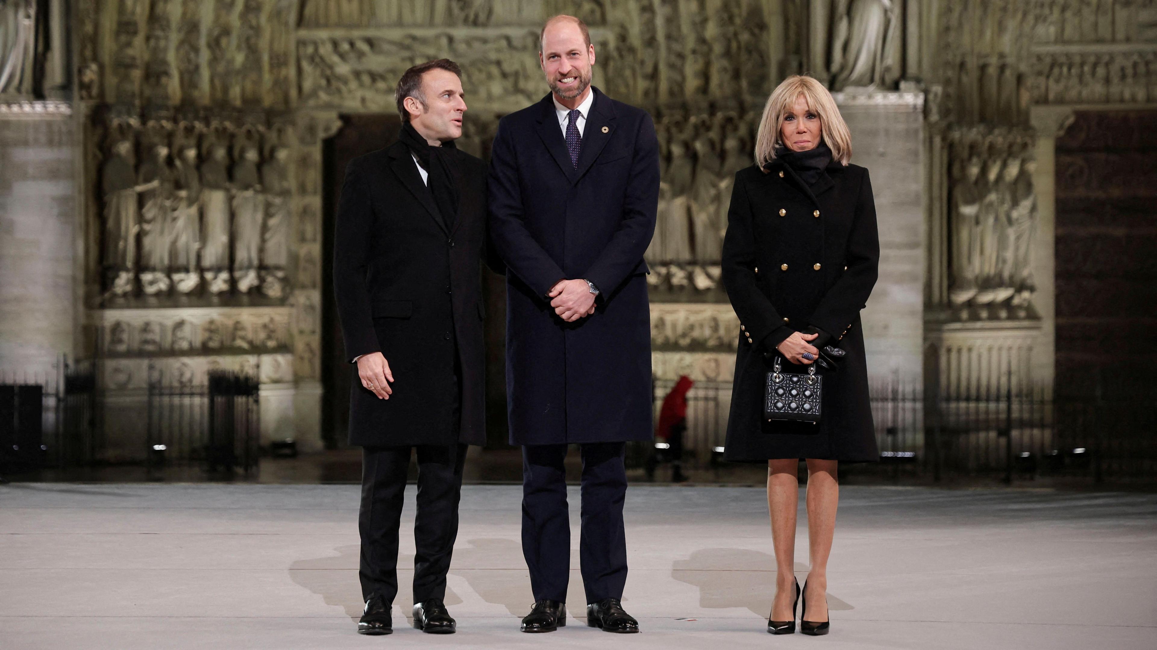 President Macron, wearing a black overcoat and scarf with a white shirt, says something to Prince William, who is also wearing a black overcoat and a white shirt with a blue tie. Brigitte Macron, wearing a black coat, black scarf and beaded black bag looks on beside them as they stand outside the Notre Dame 