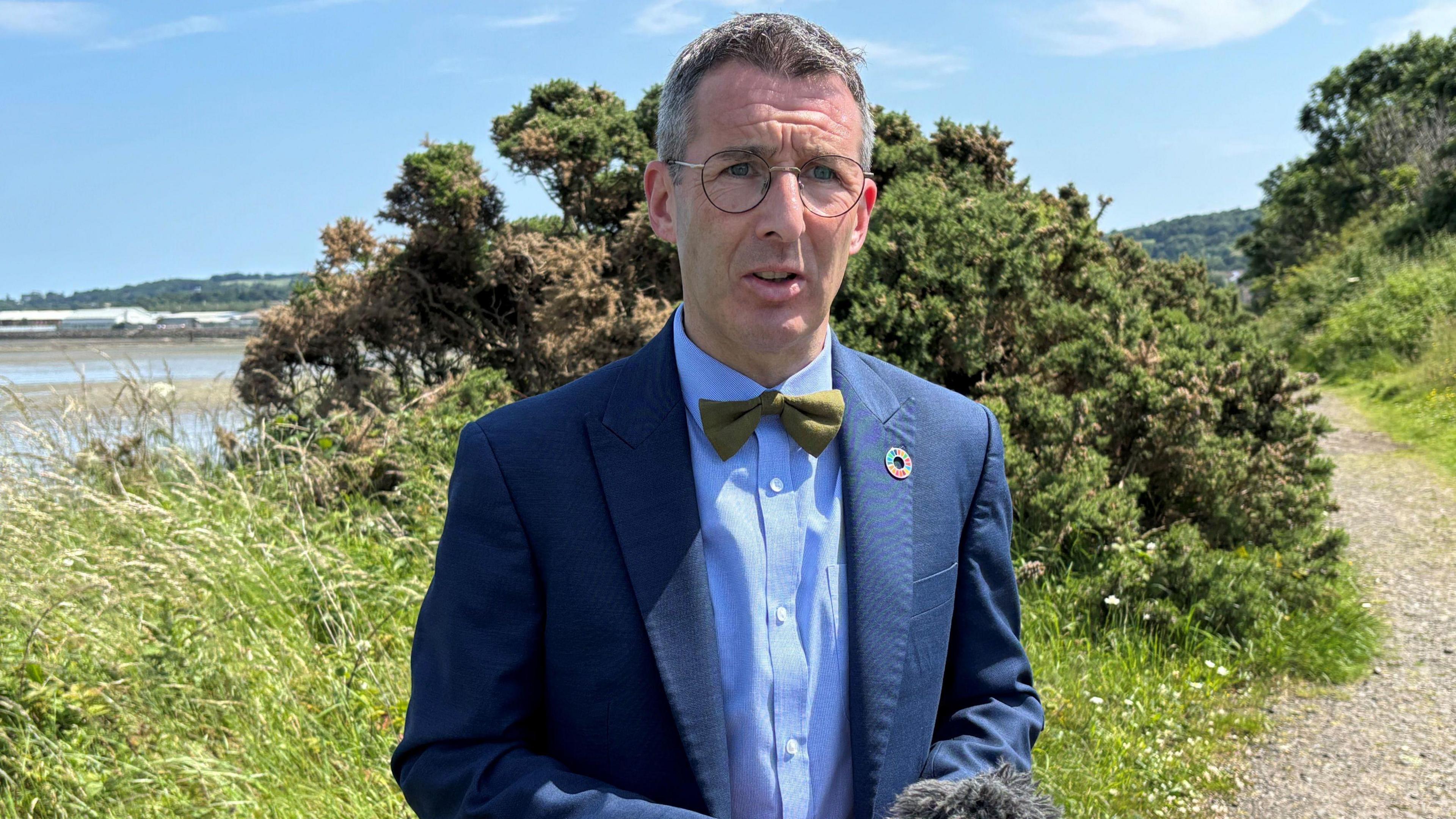 A man with short grey-brown hair wears round spectacles. He wears a navy suit, blue shirt and green bow tie. Behind him is a large green bush which sits on a plot of grass. behind that is a large body of clear water. A path runs up the side of the water.