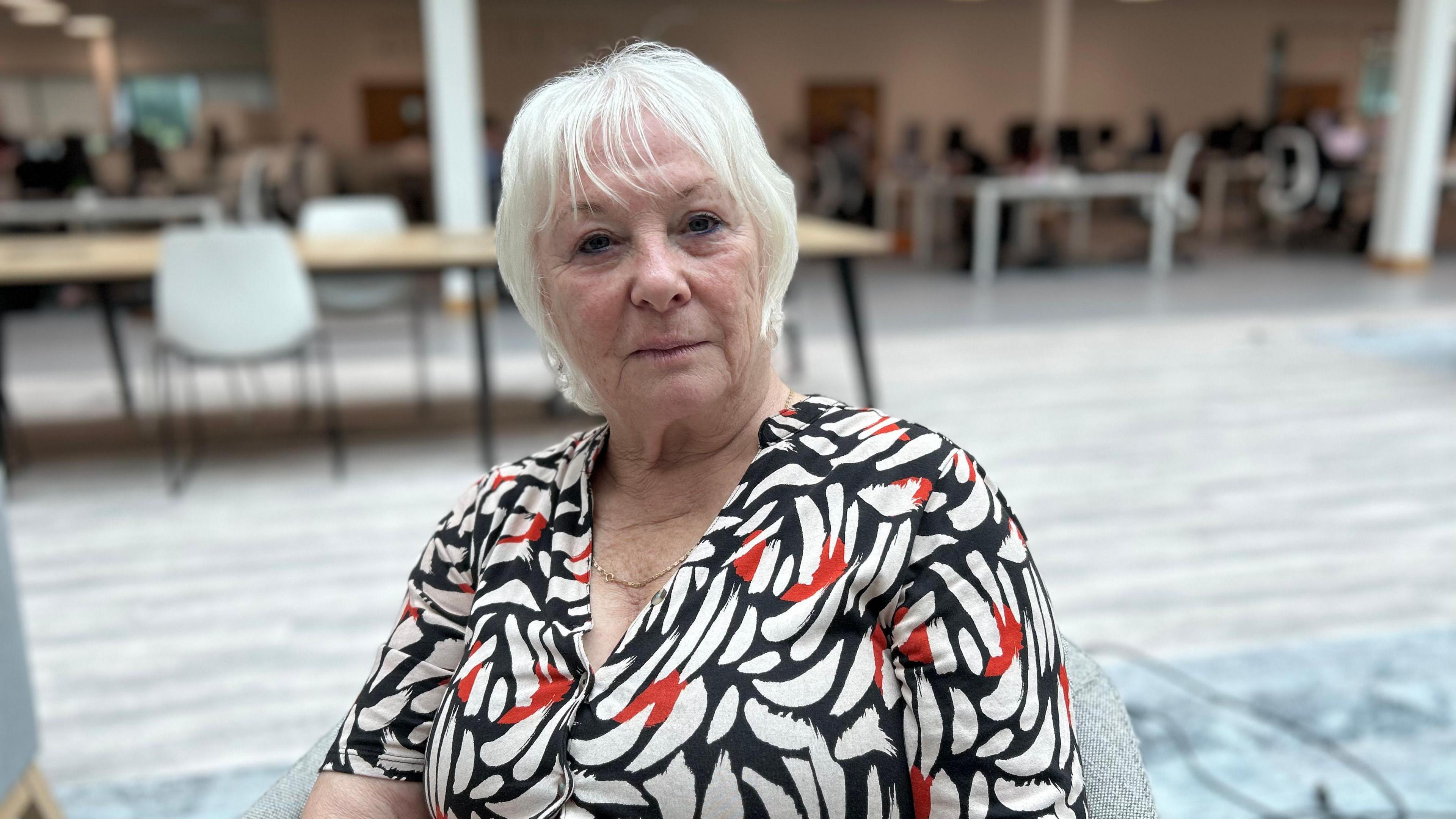 Danielle Stone in a white, red and navy patterned dress sits in a grey chair looking at the camera with lots of tables and chairs in the background