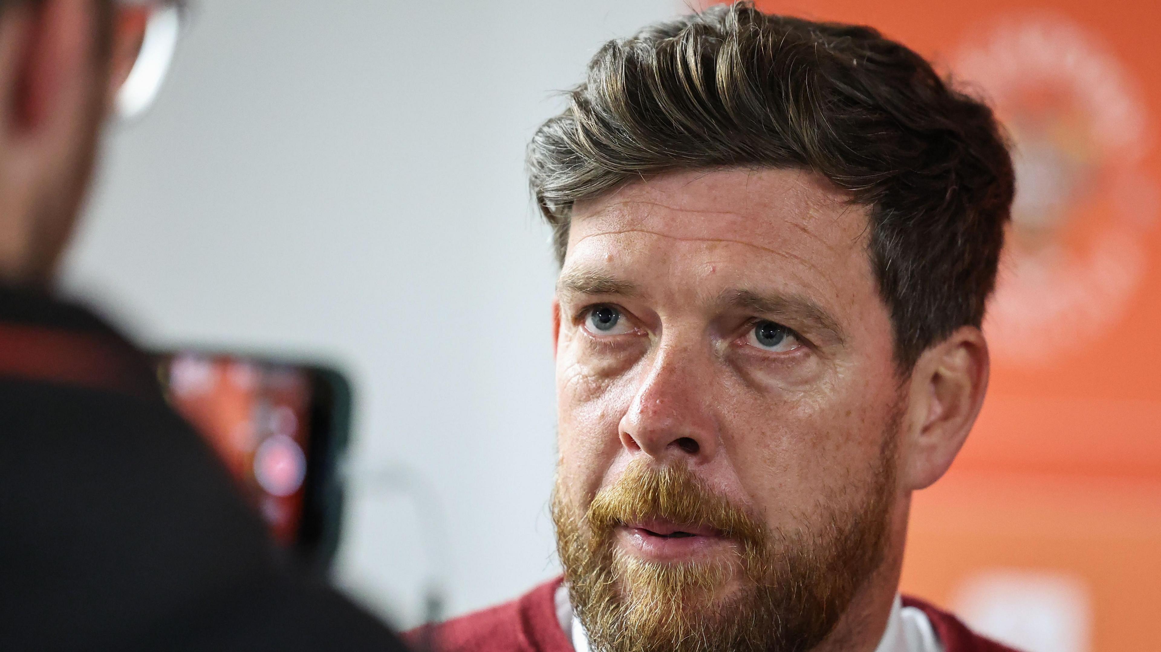 A photo of a white man with dark hair and a beard, he is wearing a white top with a red jumper. To his left is the man holding a phone 