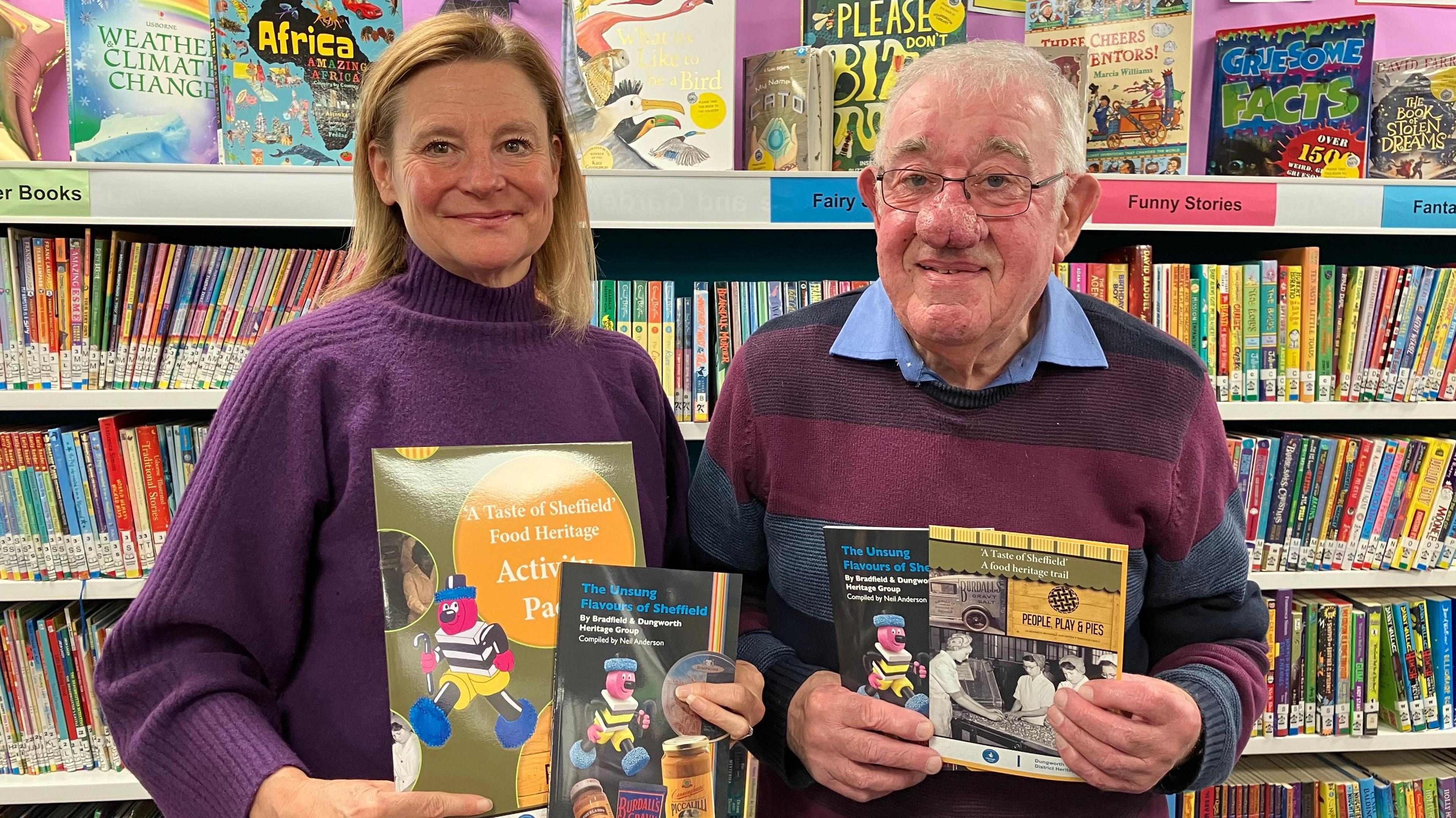 A woman and man hold up the book, with a black cover and blue title, and smile at the camera. They are in a library with books behind them. She appears in her fifties and has shoulder length blonde hair and a purple jumper. He appears in his seventies, with glasses, white hair and a blue shirt under a striped black and maroon sweater.