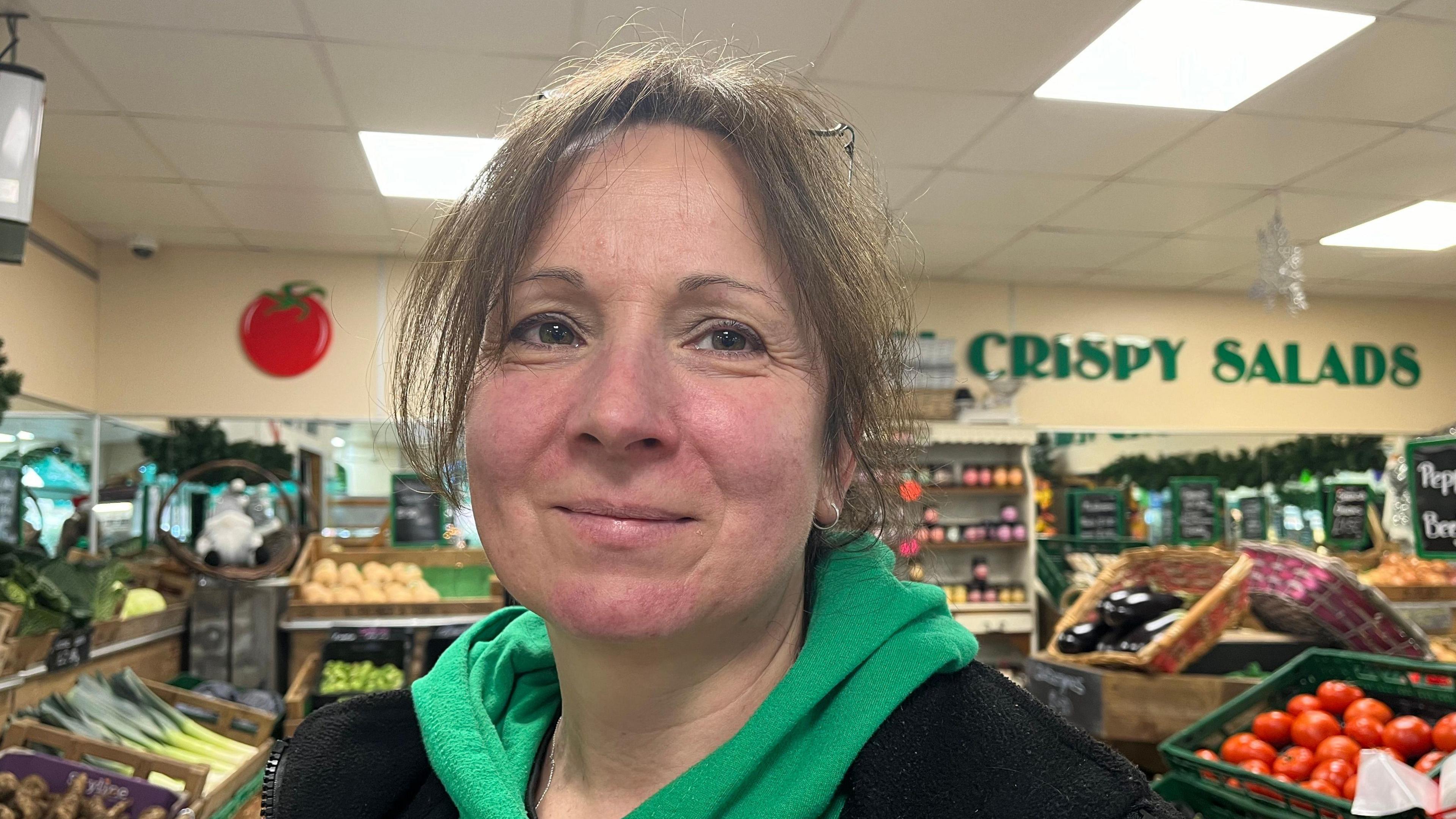 Trudie looking into the camera, she's wearing a green hoodie with a black fleece over the top. Behind her you can see rows of fresh fruit and vegetables, and a sign on the wall reads 'crispy salads'.