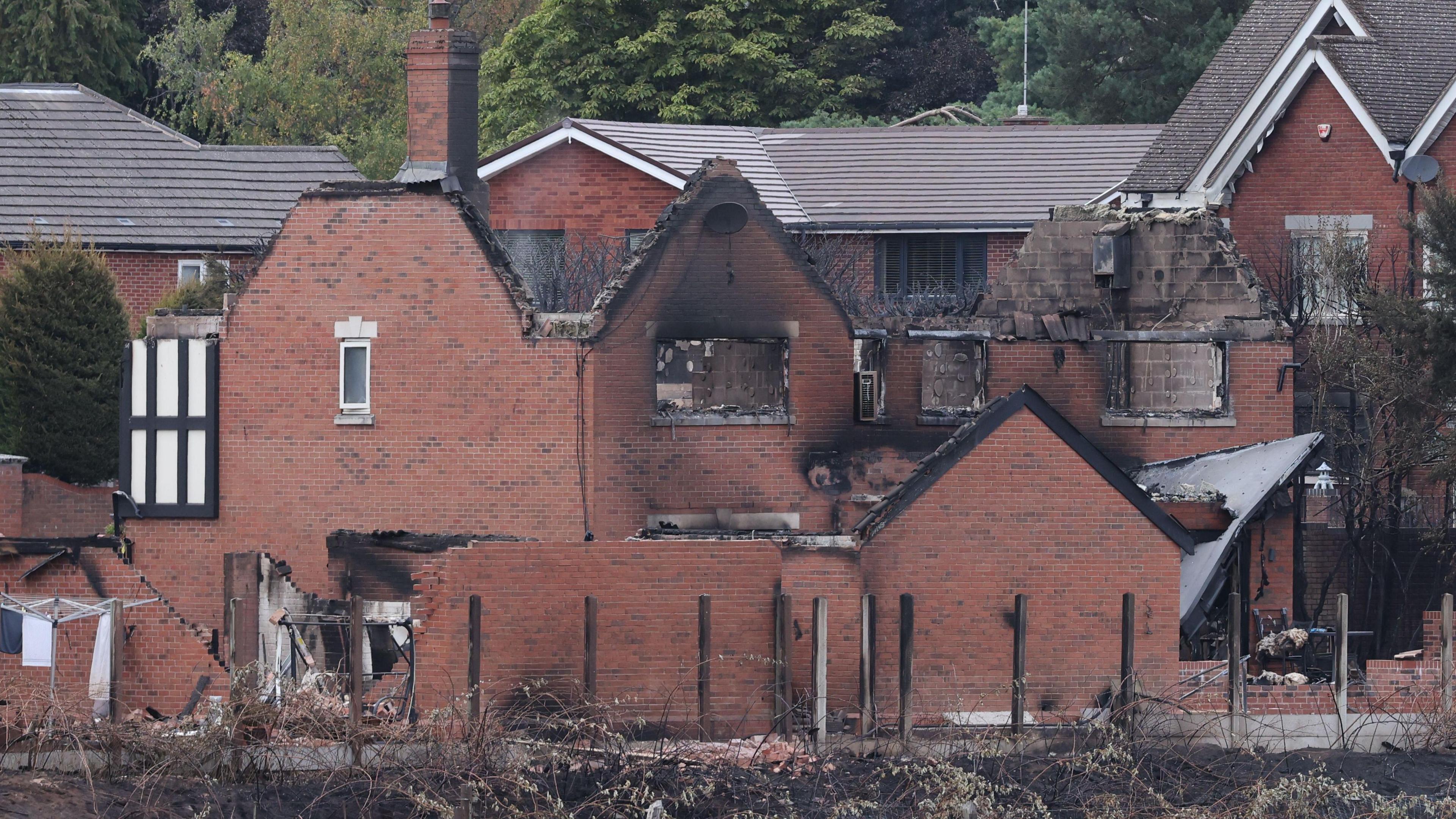 A red brick house left in ruins from fire, with its roof burned off, and black singes around the edges of the ruins. Burnt shrubbery can be seen in front of it, with houses behind still in tact.