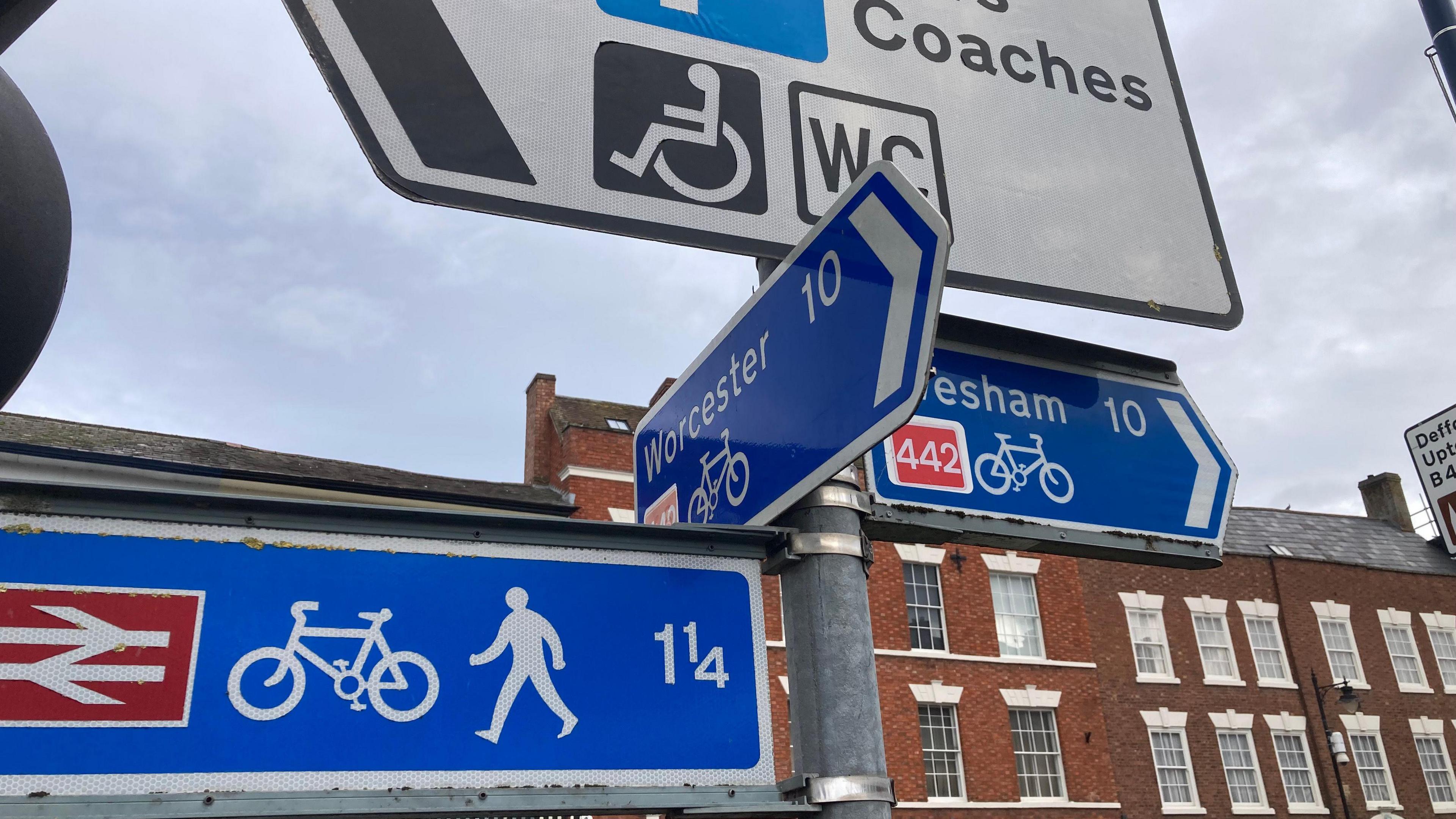 A street sign, showing a left turn towards Worcester and a right turn towards Evesham. There are some buildings in the background. 