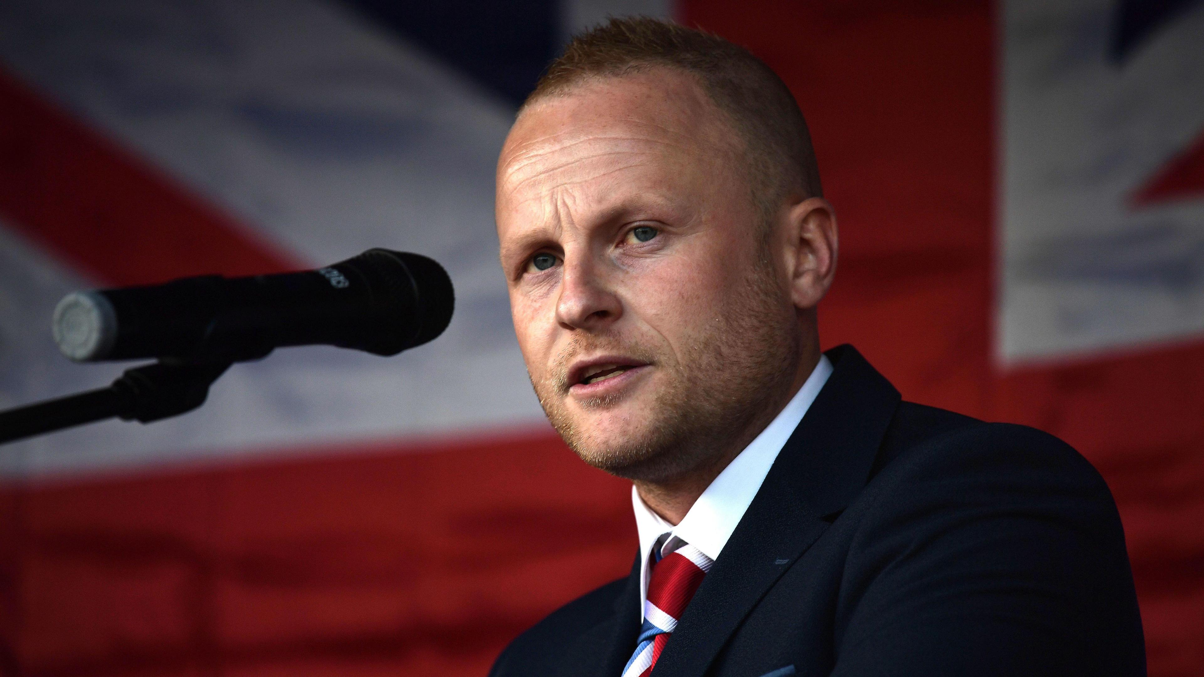 Jamie Bryson speaking into a microphone at a 2021 protest against the post-Brexit Irish Sea trade border. He has short blonde hair and is wearing a navy suit and union flag tie. There is a union flag in the background.