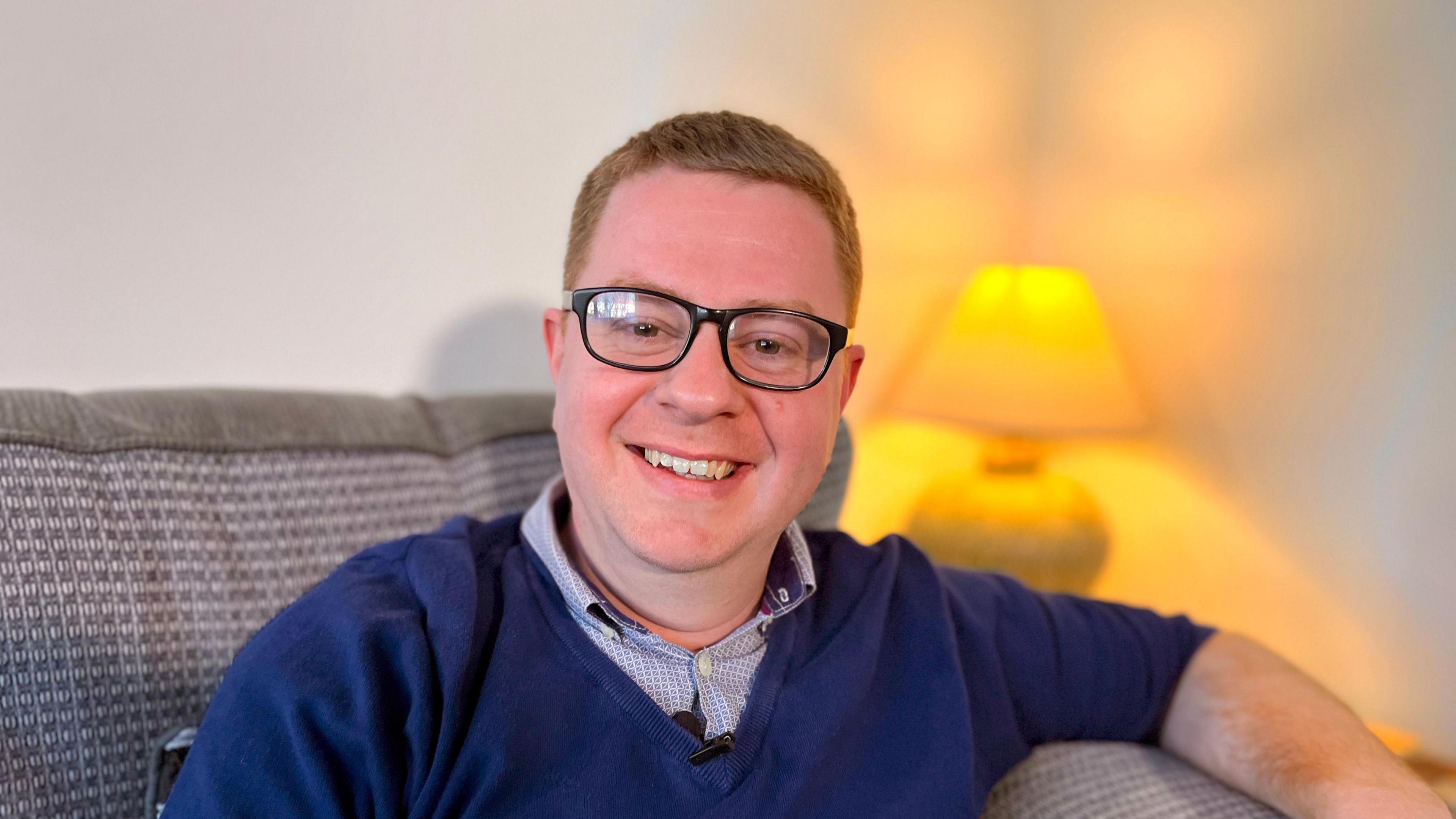 A smiling sandy haired man with dark glasses. He is wearing a navy blue V-neck sweater over a patterned shirt. He is sitting on a sofa with a lamp in the background. 