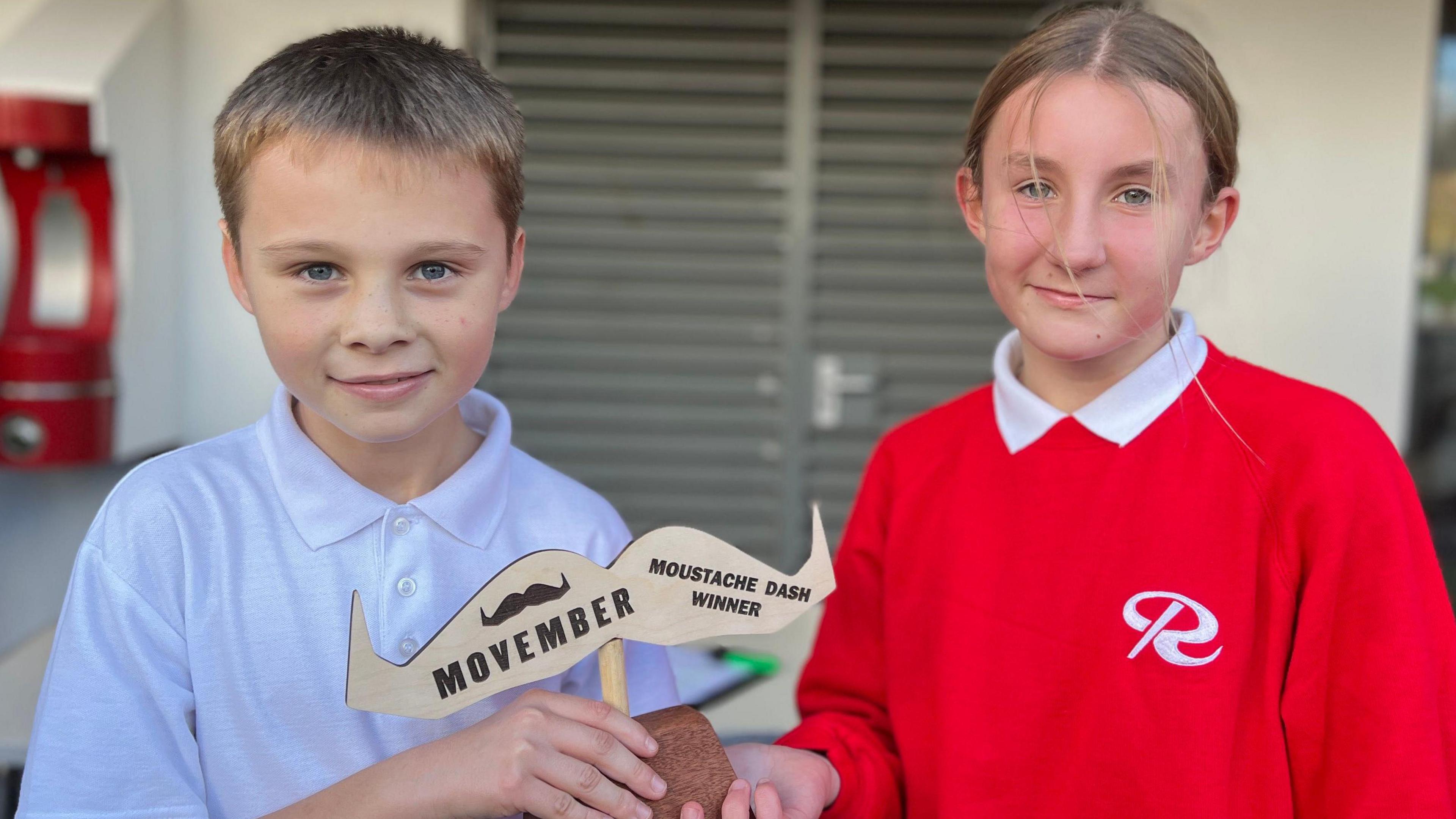 Stanley and Emily are standing outside near a building with gray shutters. Stanley is wearing a white polo shirt and holding a wooden trophy shaped like a mustache with the text “MOVEMBER” and “MOUSTACHE DASH WINNER.” Emily is wearing a bright red sweatshirt with a white logo on the chest. 