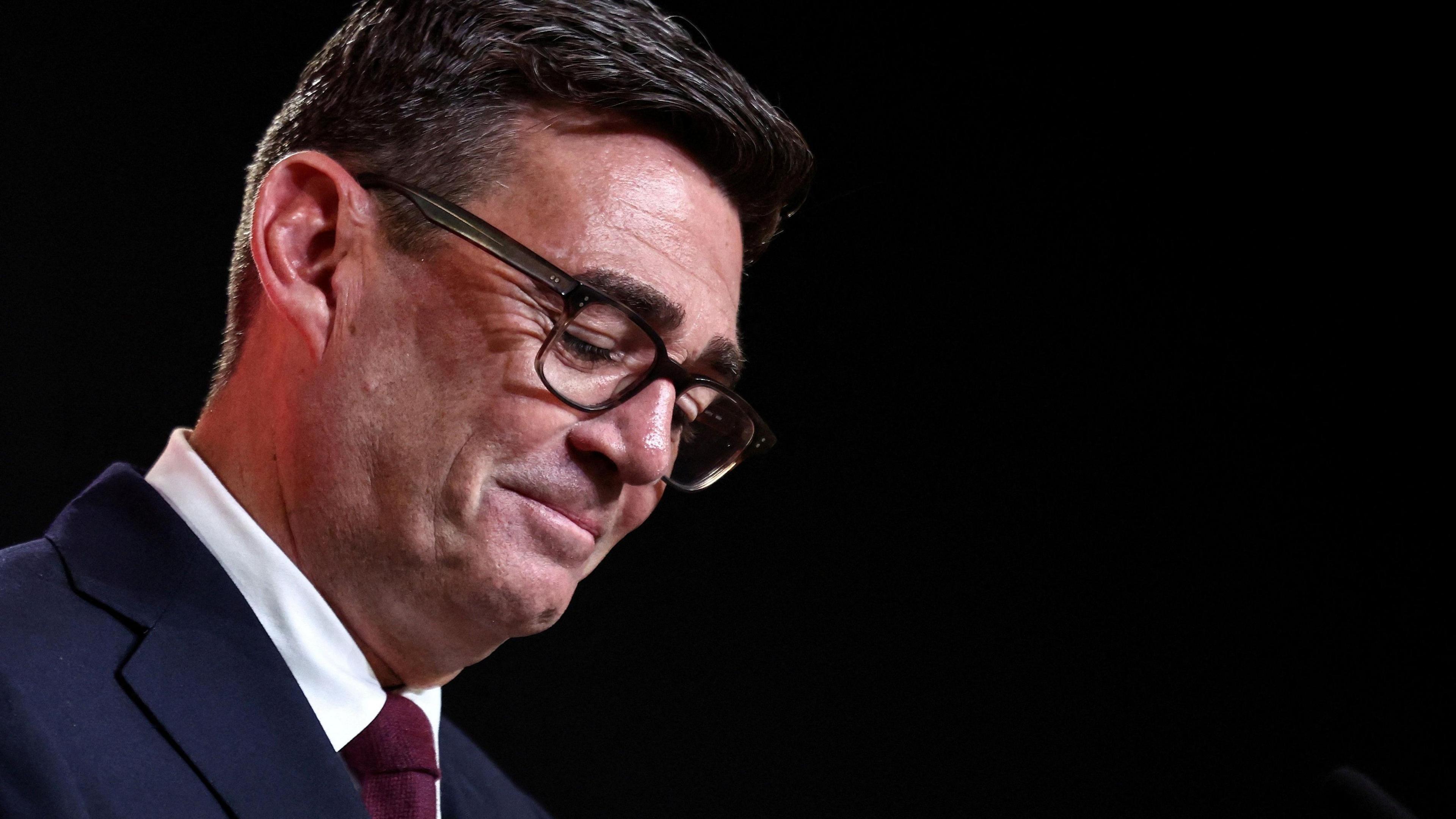 Andy Burnham, in dark blue suit with plain white shirt and solid burgundy tie, stands in front of a plain black backdrop. He is looking down, smiling ruefully, wearing dark-rimmed plastic spectacles.
