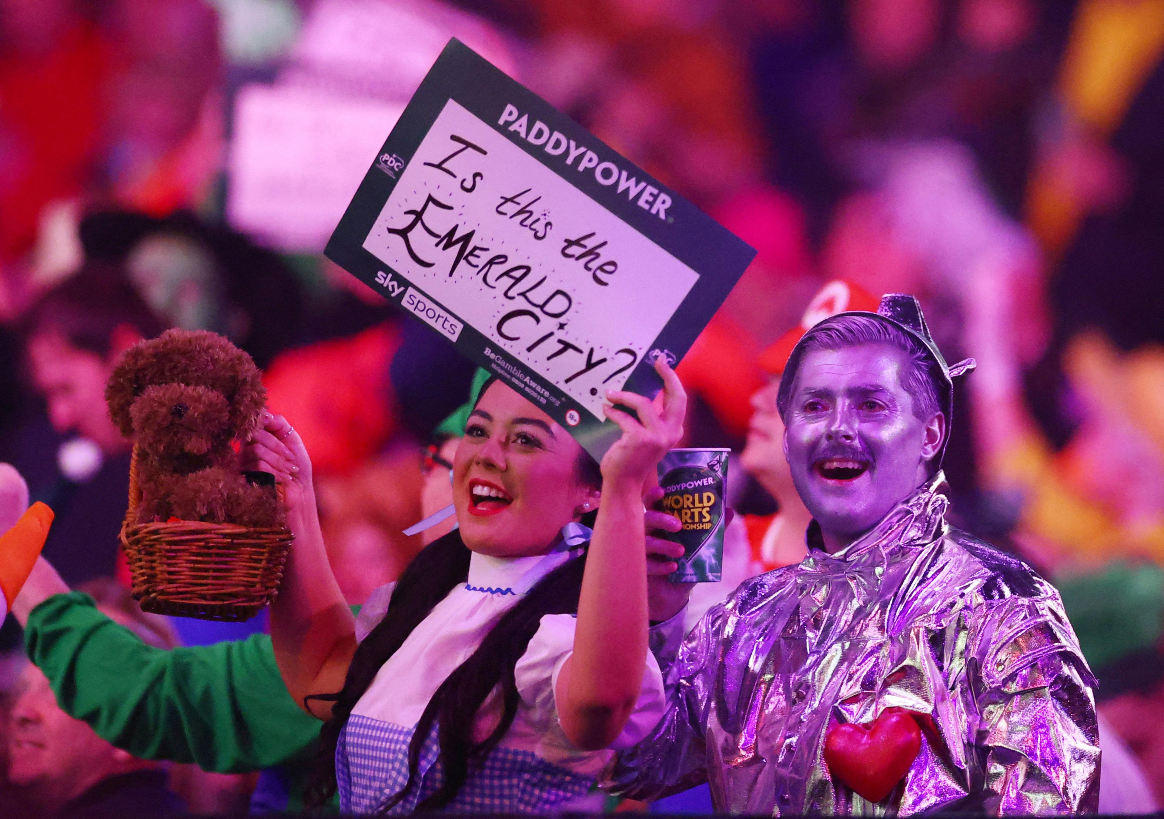 Fans in Wizard of Oz fancy dress - a woman dressed as Dorothy and a man dressed as the Tin Man - cheer at a darts match. The woman is holding a plushy dog toy and a sign saying "is this the Emerald City?" The man is holding a drink.