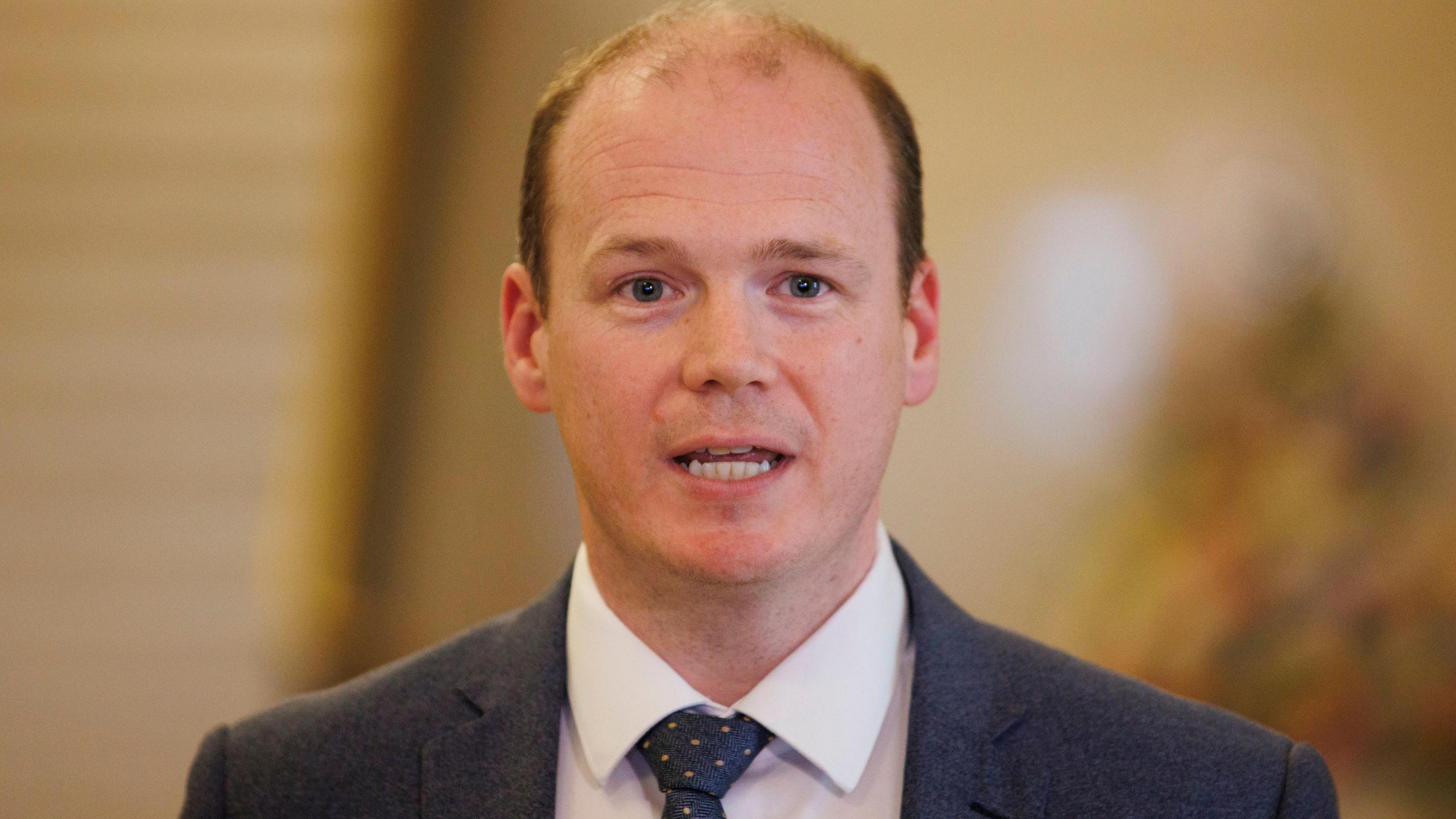 Communities Minister Gordon Lyons during a press conference at Parliament Buildings at Stormont in Belfast - he is wearing a white shirt, grey blazer and grey tie with yellow dots. He has dark hair.