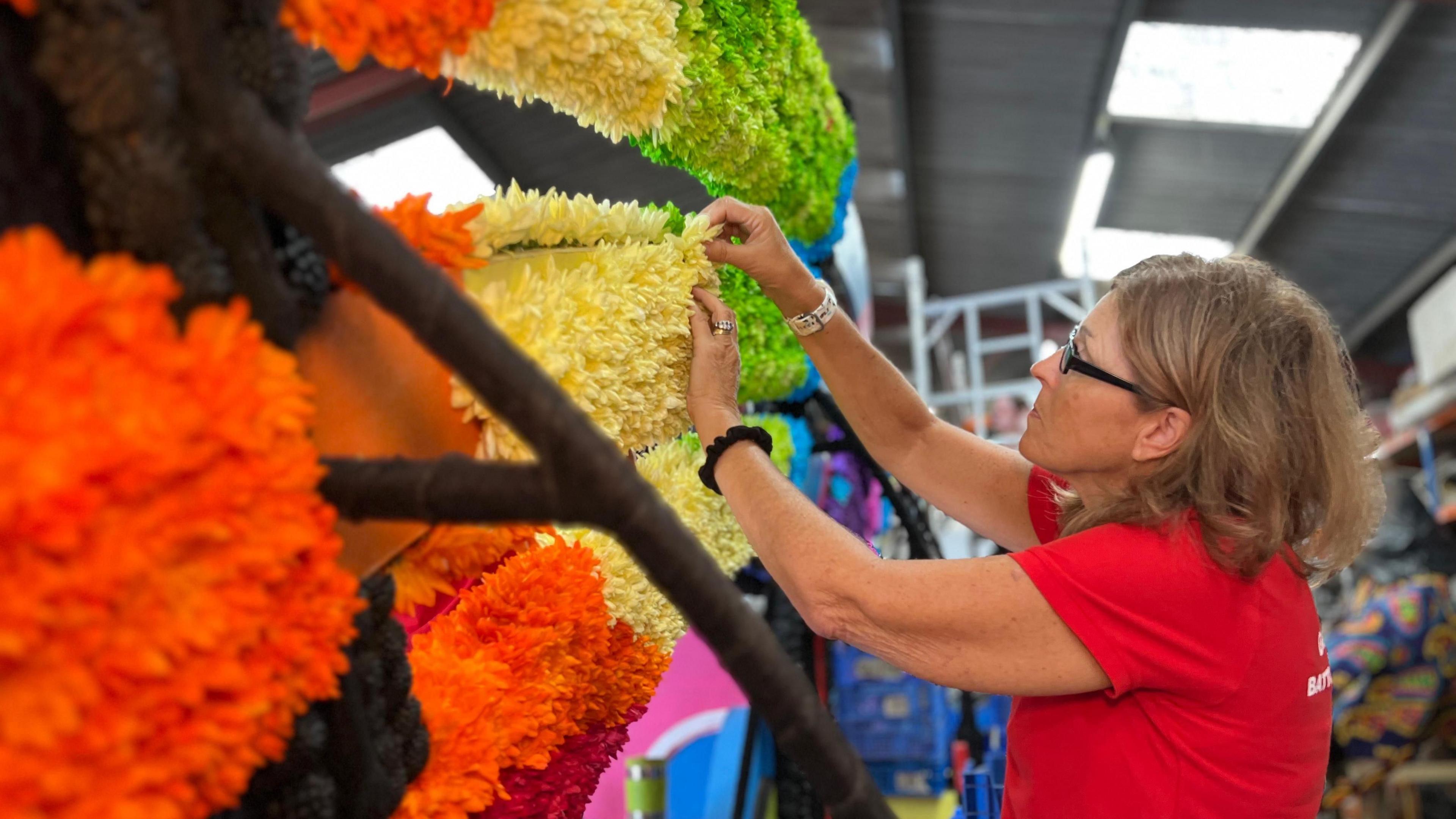 A woman in a red t shirt leaning forward and sticking a yellow flower to a floral rainbow as part of a float