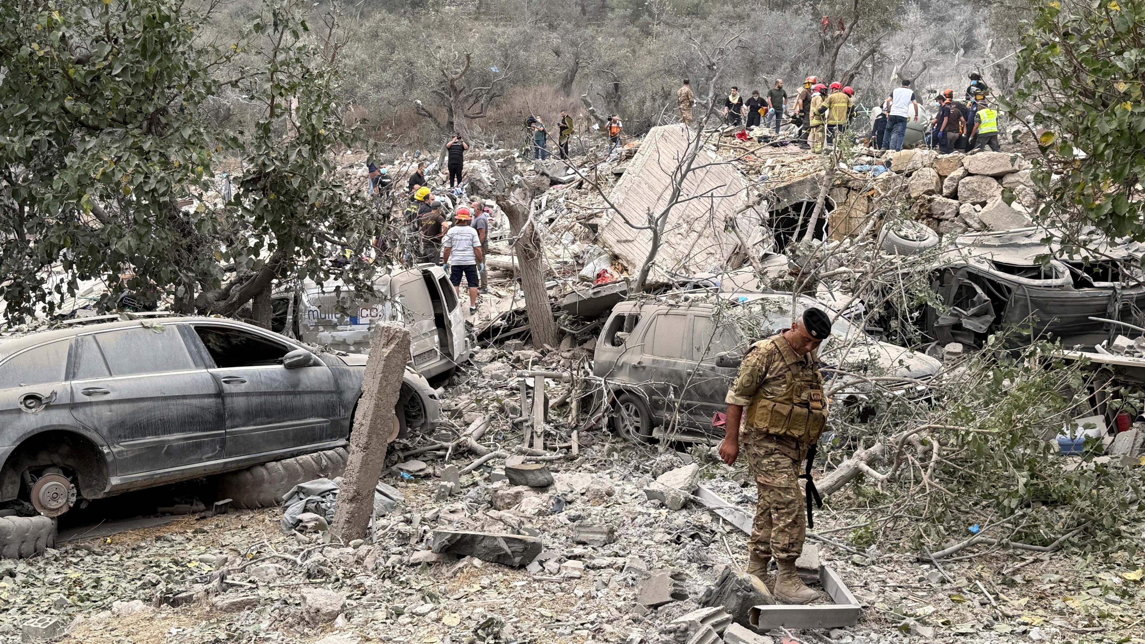 A soldier inspects the aftermath of the strike, with broken vehicles and piles of dusty detritus behind him.