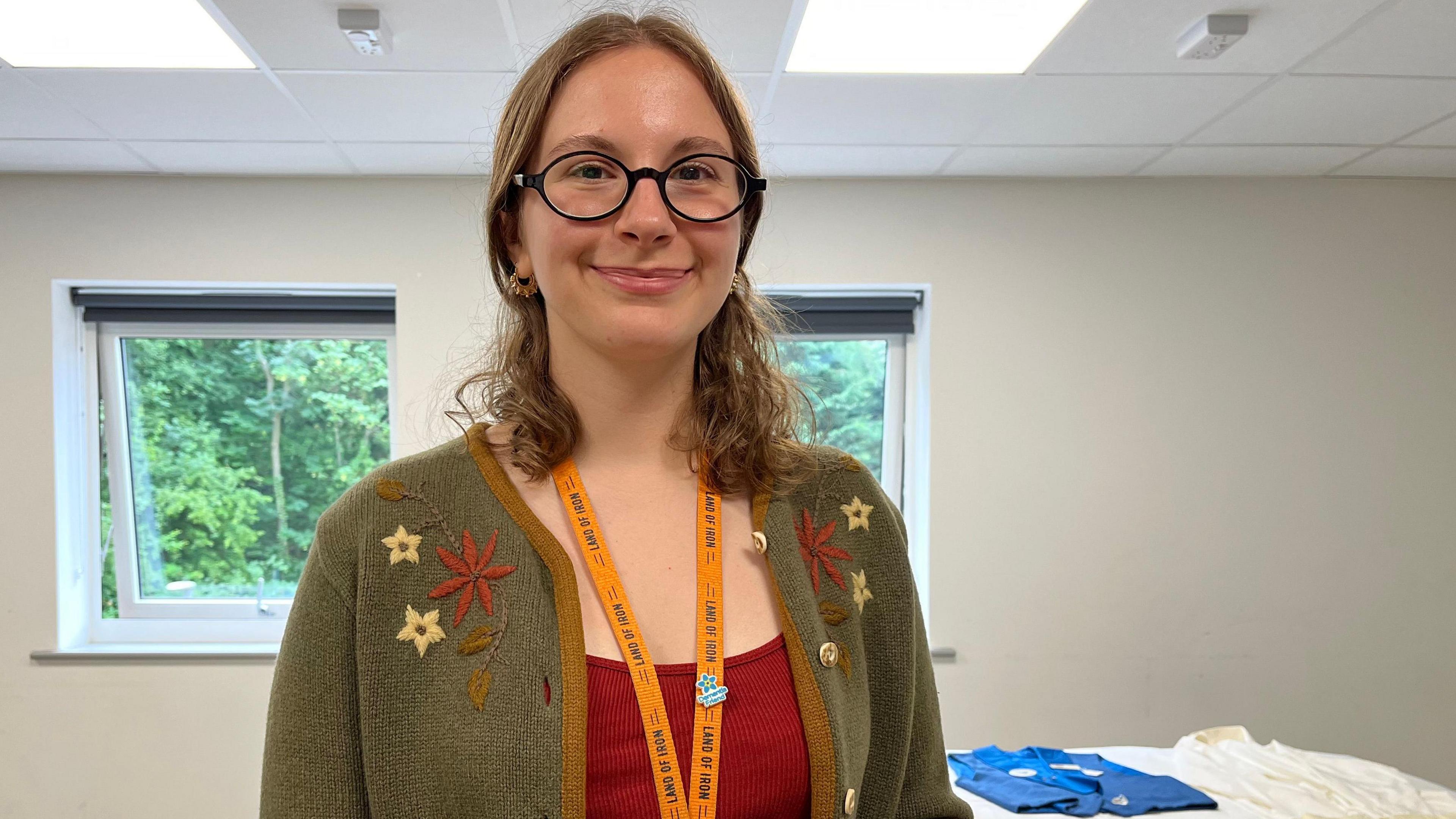 Grace Redpath is wearing an orange lanyard with a green cardigan covered in flowers. she has short brown hair and glasses and is wearing a red tank top. Behind her there is memorabilia including sword dance vests and photographs.