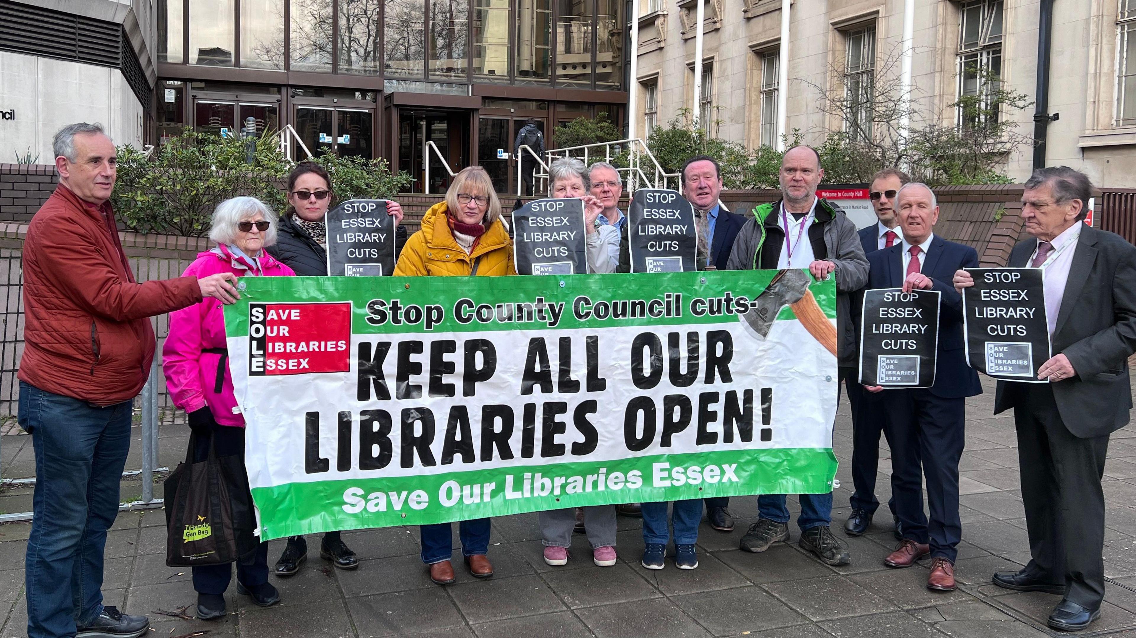 About a dozen people standing outside an office building. Many of them are holding signs saying "STOP ESSEX LIBRARY CUTS" in white lettering on a black background. In front of them is a white and green banner that says: "Stop County Council cuts - KEEP ALL OUR LIBRARIES OPEN! Save Our Libraries Essex."