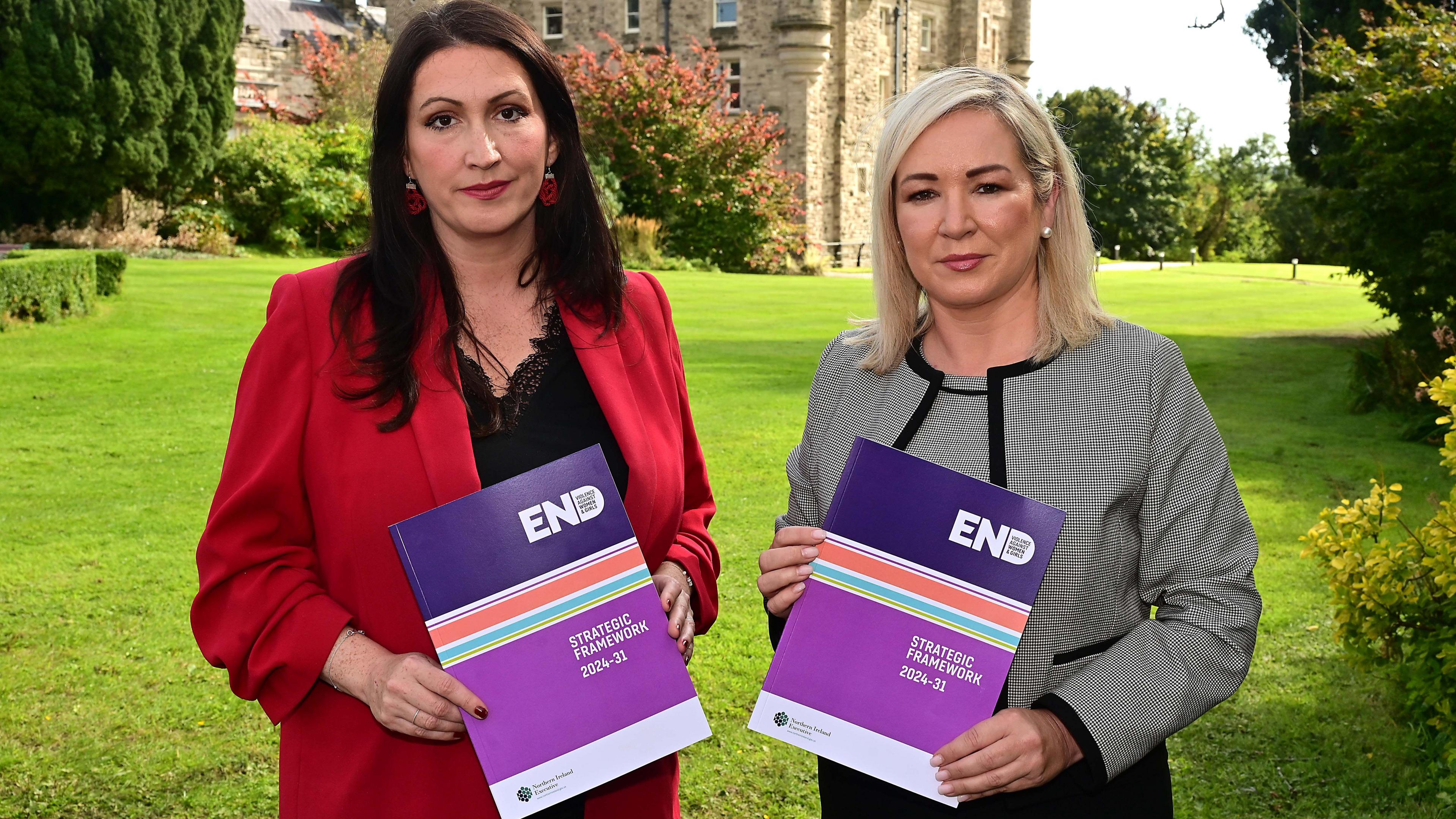 Emma Little-Pengelly and Michelle O'Neill holding up copies of the strategy. The backdrop is Stormont Castle and the pair are standing on the lawn. Emma Little-Pengelly is wearing a red blazer, while Michelle O'Neill is wearing a black and grey outfit