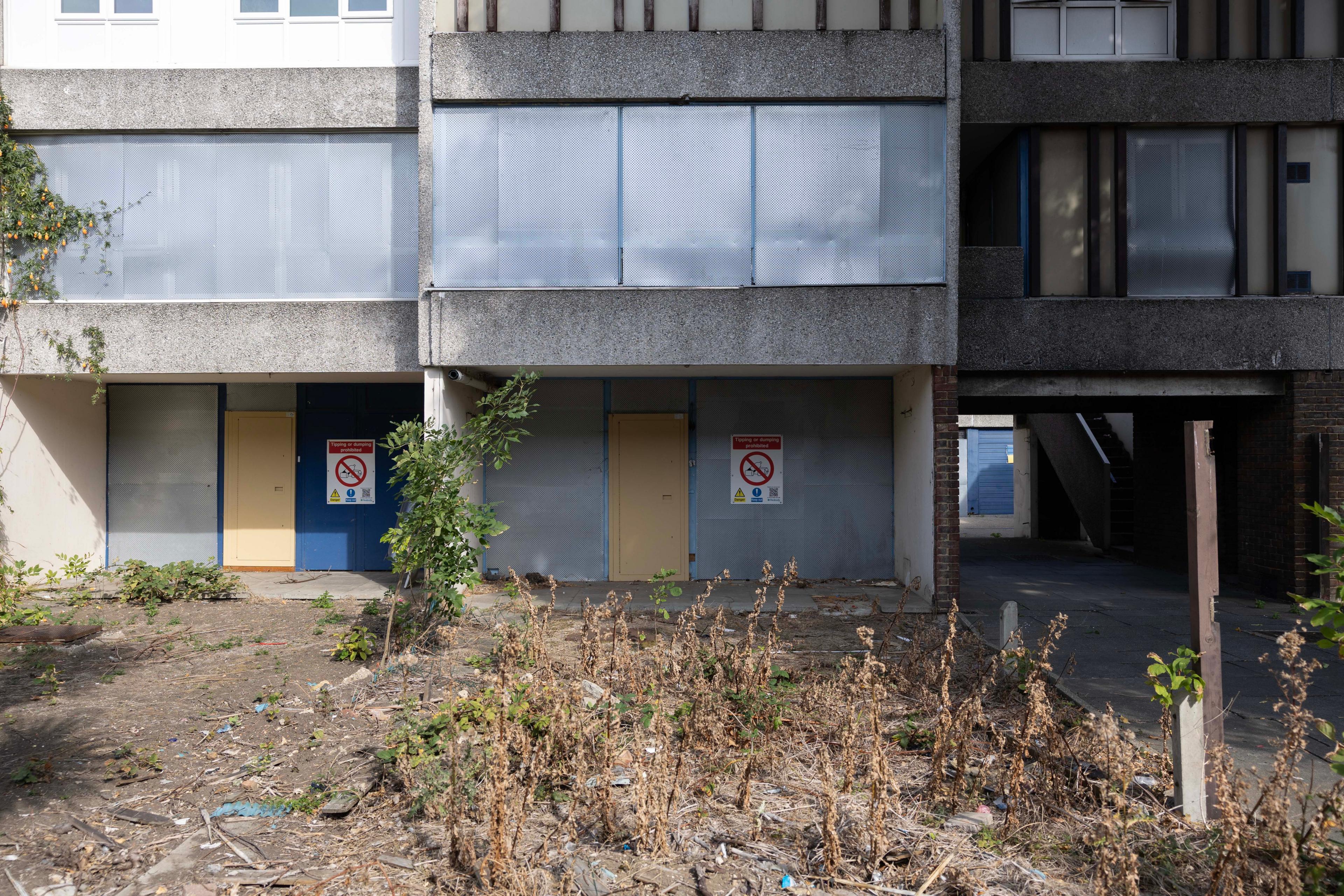 Derelict housing estate shows overgrown weeds with boarded up entrances and windows