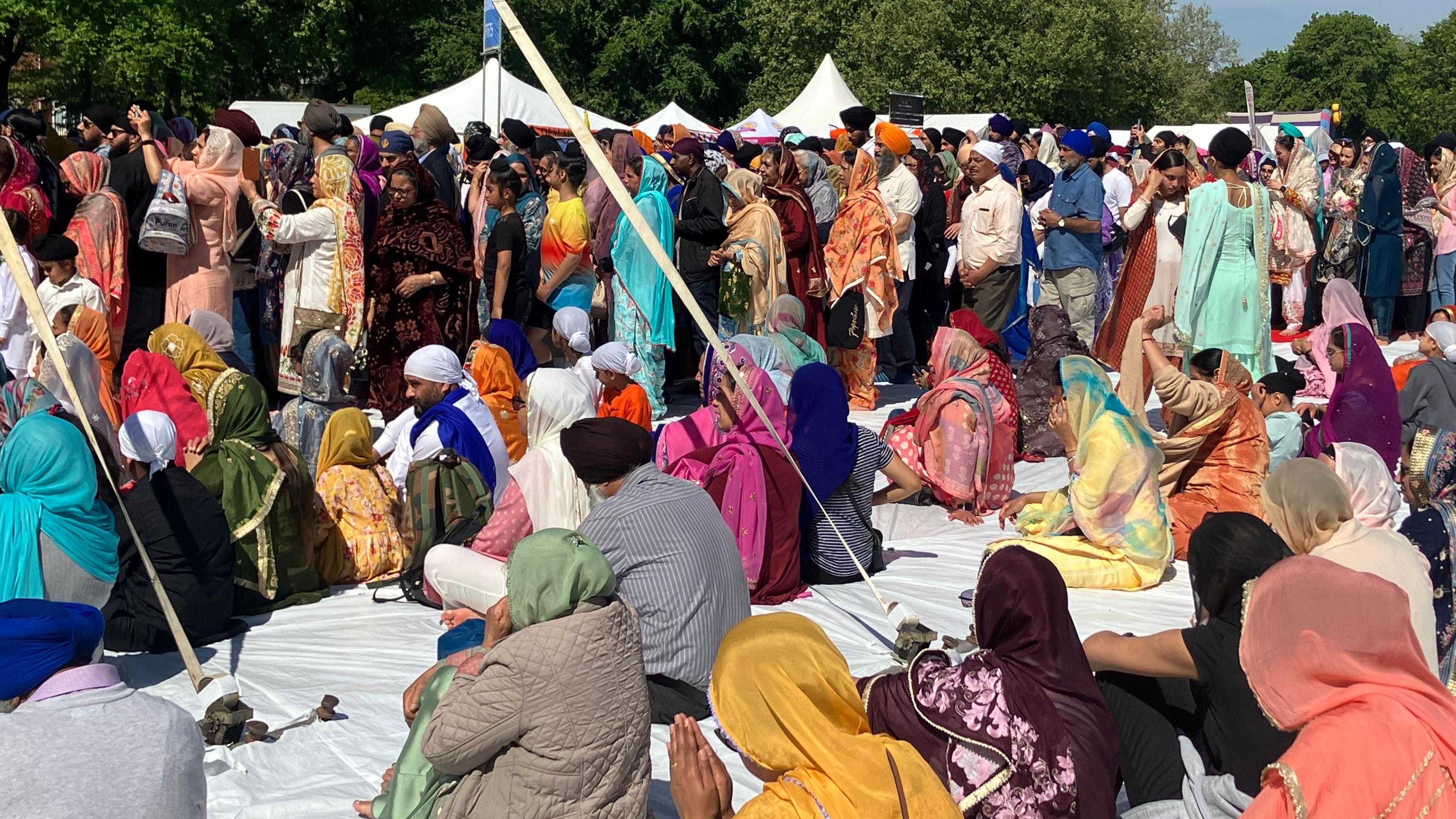 A group of more than 100 people dressed in traditional south Asian attire. The people closest to the camera are seated on a white sheet on the ground while people are standing in the background.