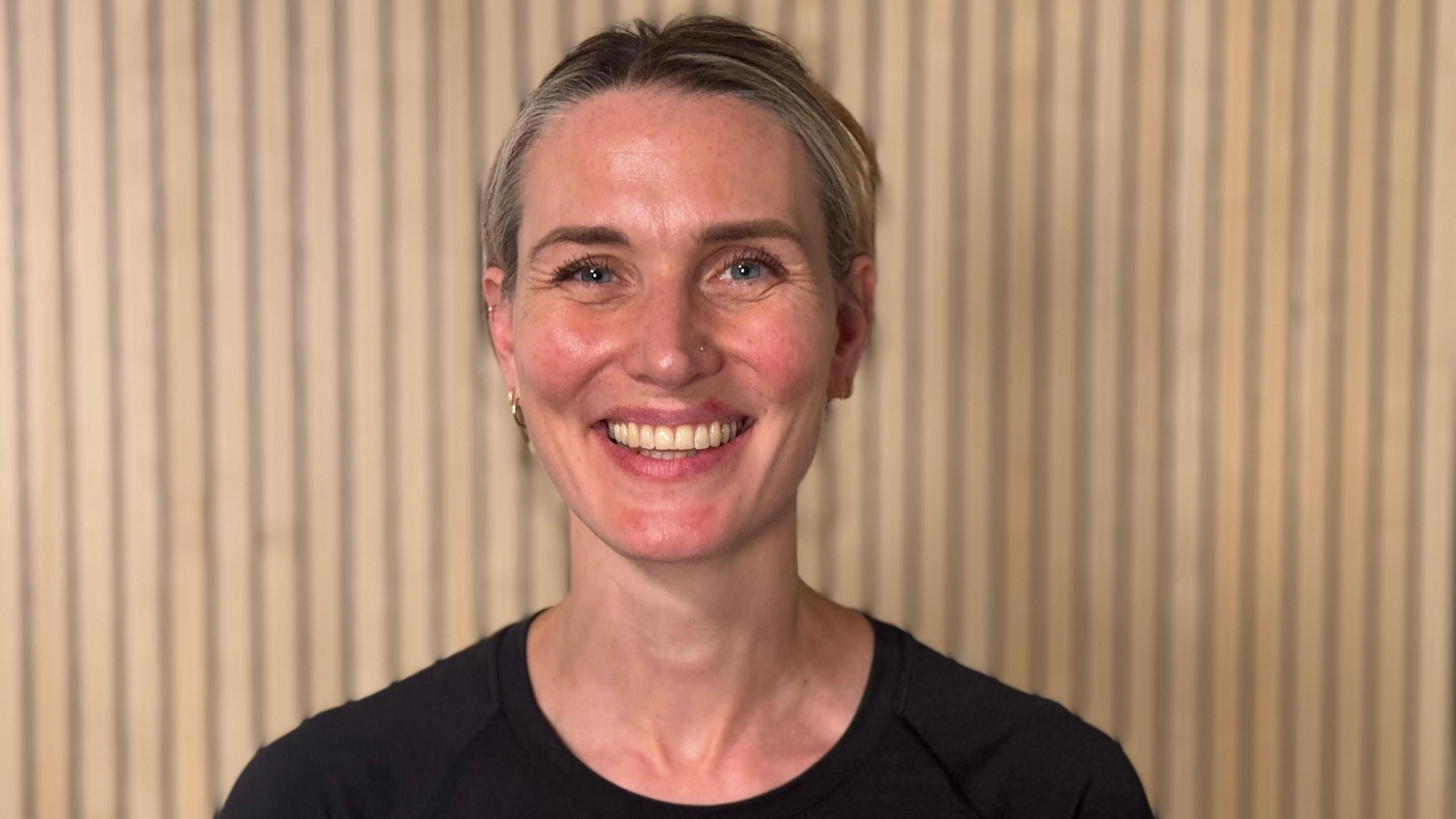 Physiotherapist Kirtsy Henderson stands in front of a wooden-clad wall in the studio where she records her exercise videos. She is wearing a black t-shirt, her hair is tied back and she is smiling.