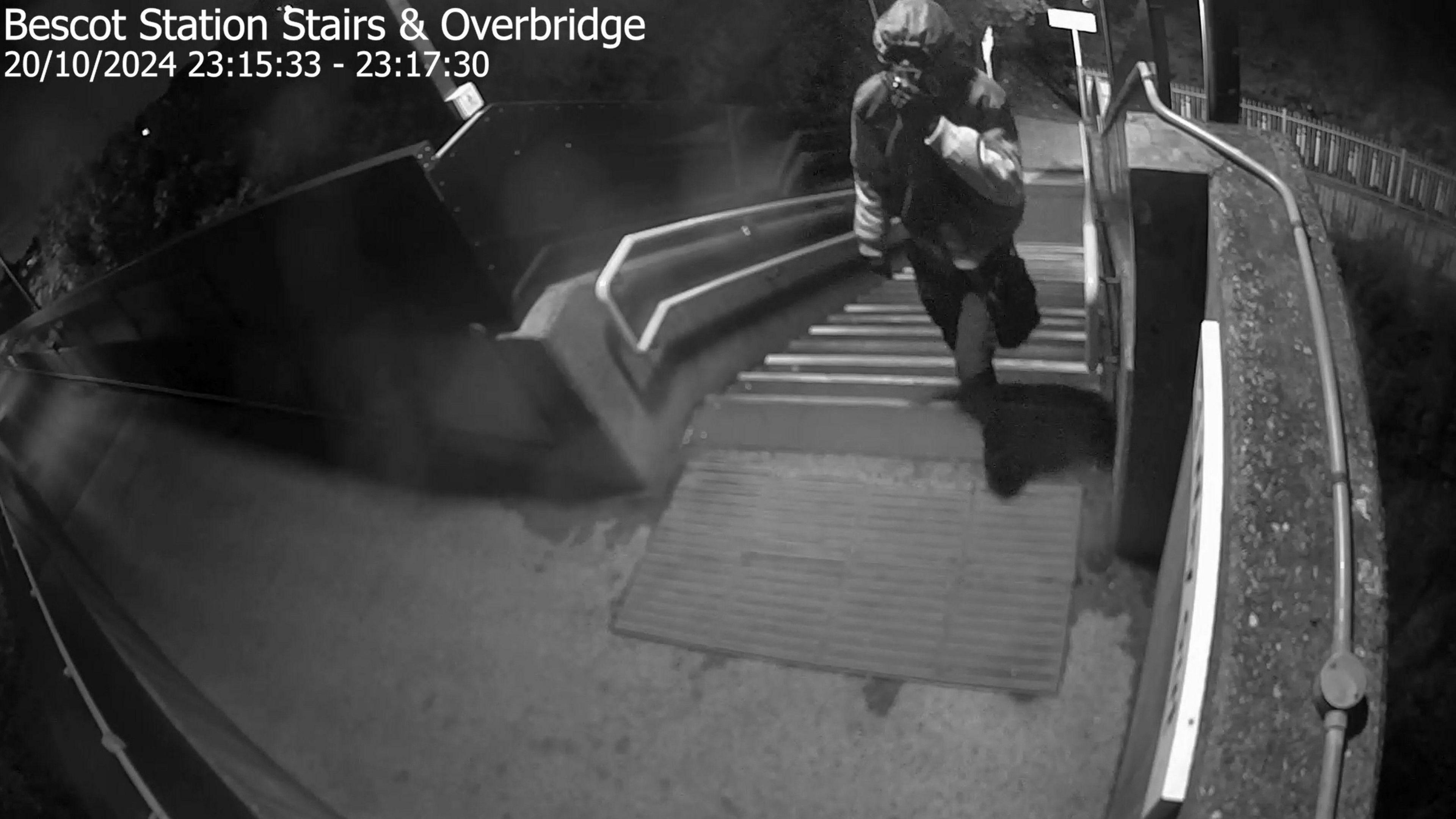 Black and white CCTV footage shows a person with their hood up, walking up some steps onto a railway bridge at night