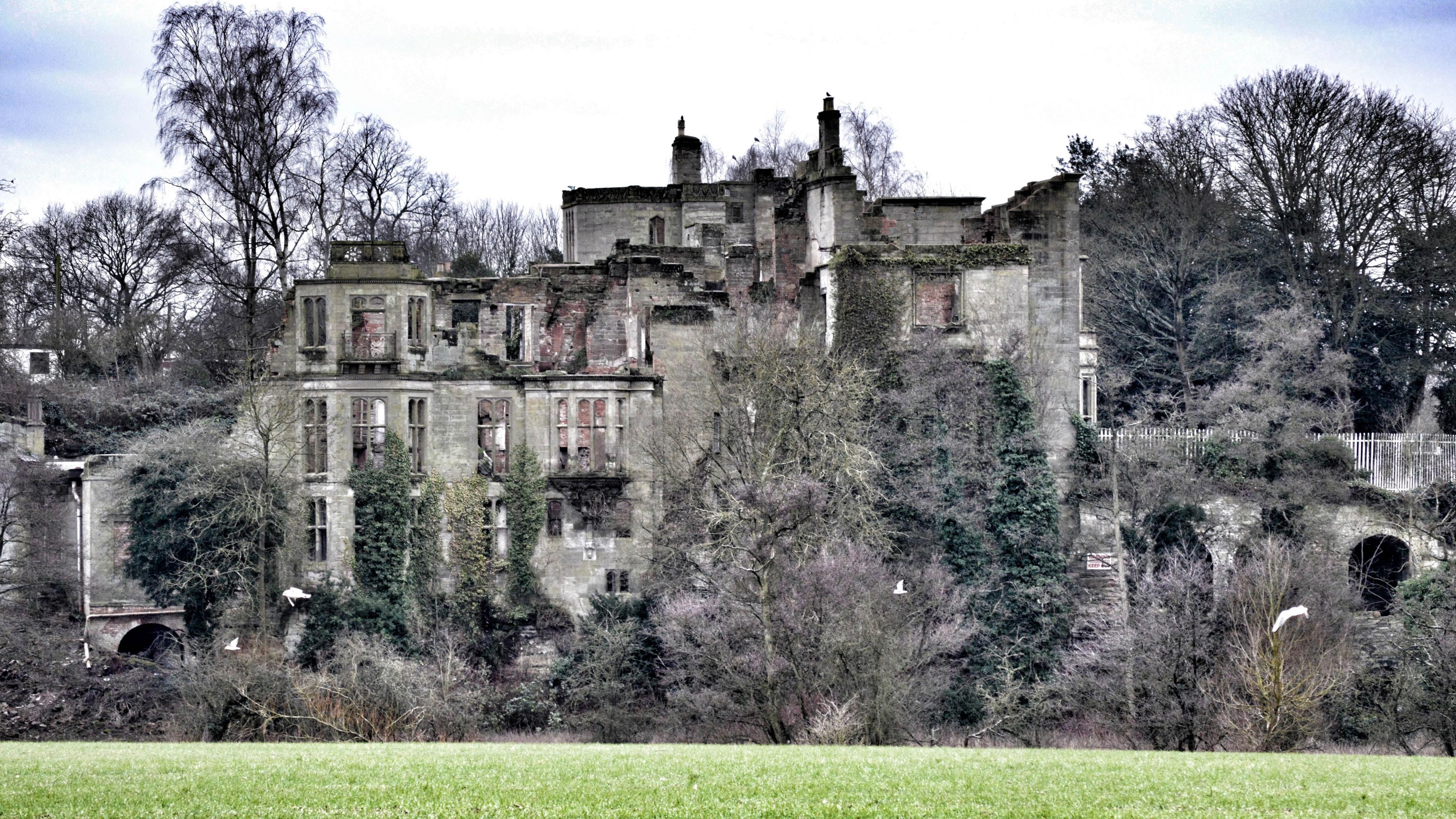 A semi derelict stately home with green trees and a green field in the foreground