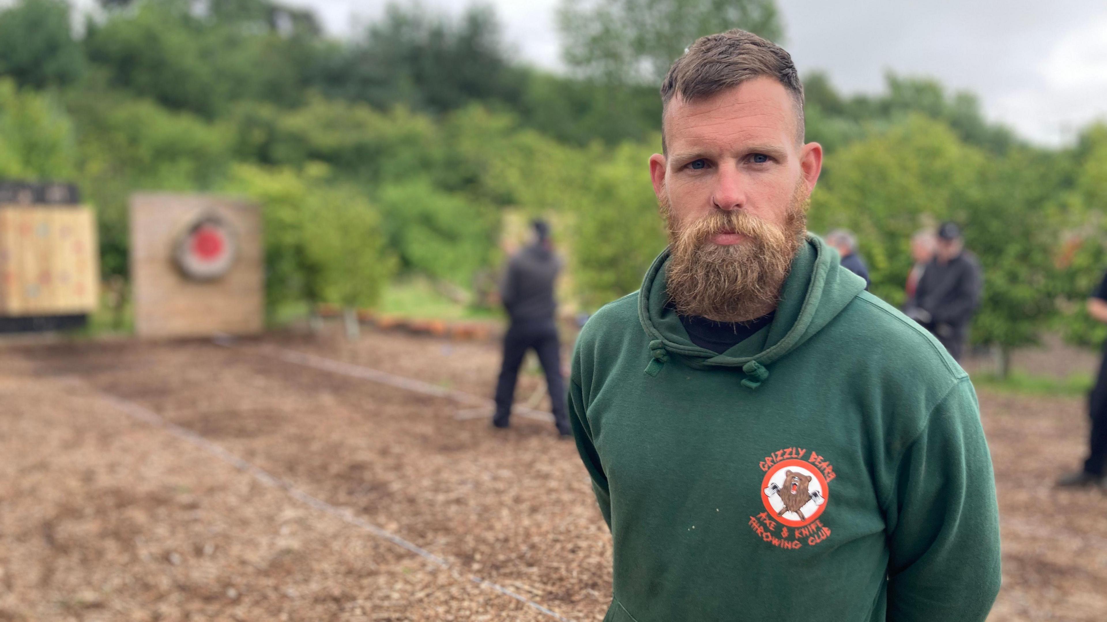 Spencer Kennedy sports a bushy, light brown beard and wears a green hooded top. In the background is a brown pitch where a person stands throwing an axe towards a red target on a wooden wall.