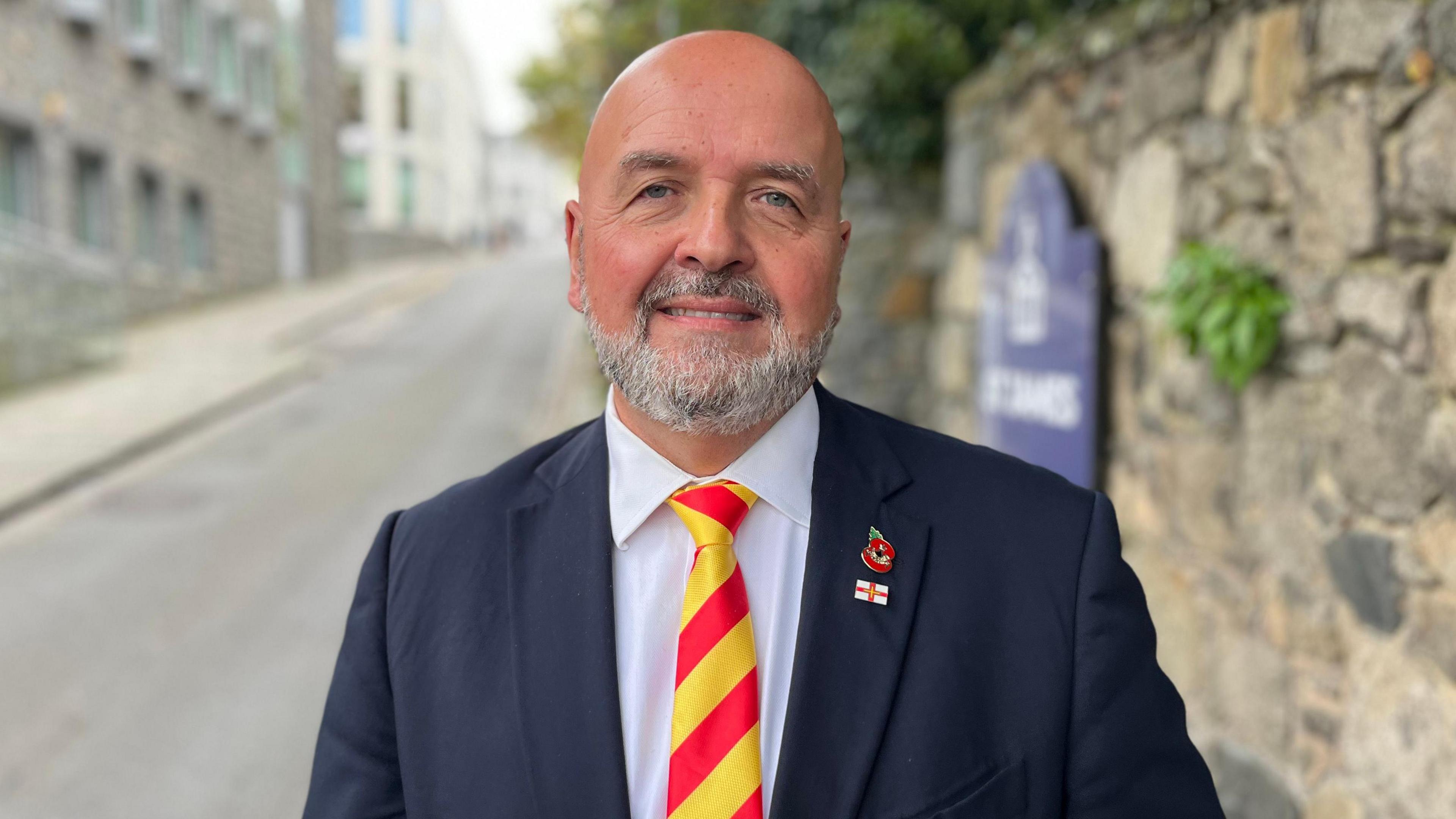 A picture of a man with no hair and a grey beard. He is wearing a navy suit jacket with a white shirt and red and yellow striped tie.