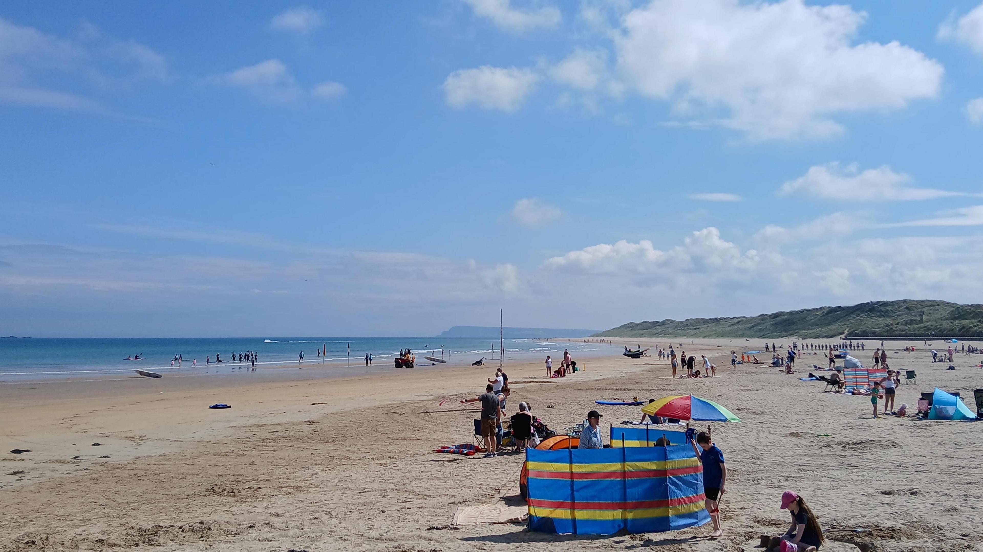 A beach with colourful windbreakers and people seemingly enjoying the conditions. Lots of sand and sea in the distance with some blue skies above