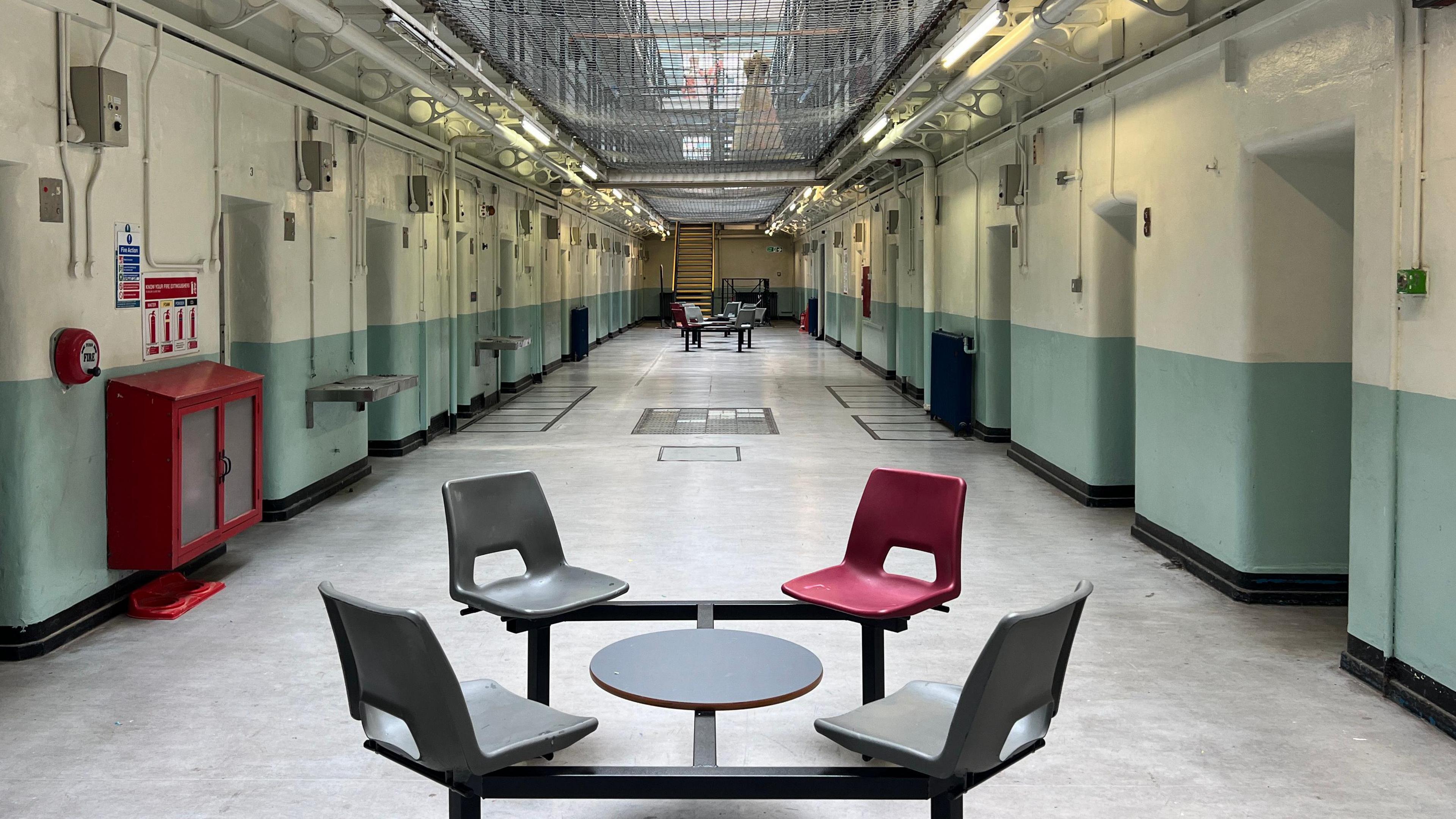 A set of four chairs sit inside a prison building with green and white walls and a caged walkway sits above it. 