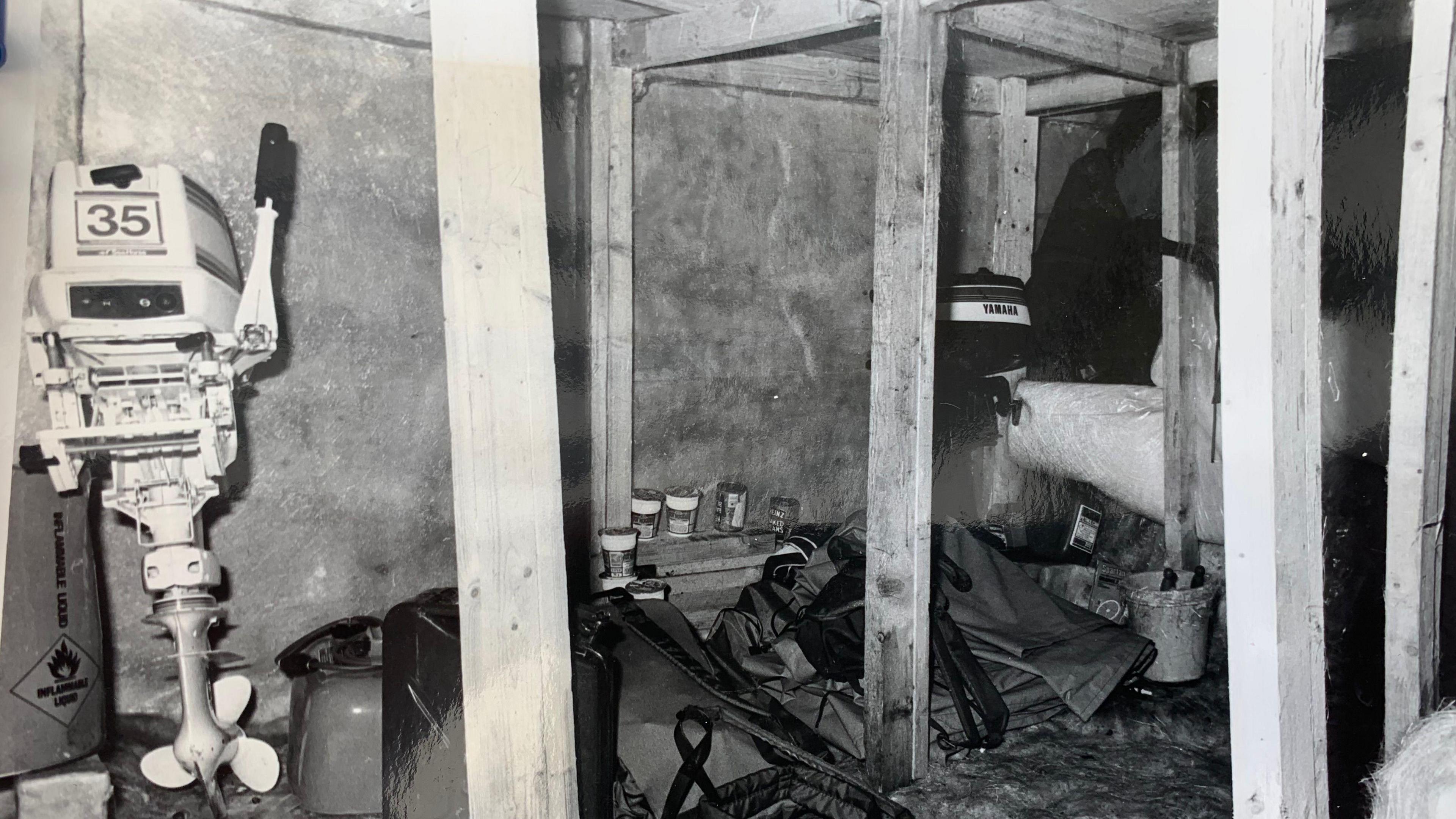 A black and white photo of an underground bunker with large timber beams and covered in fibre glass. Inside the bunker are motorboat engines and other boating equipment.