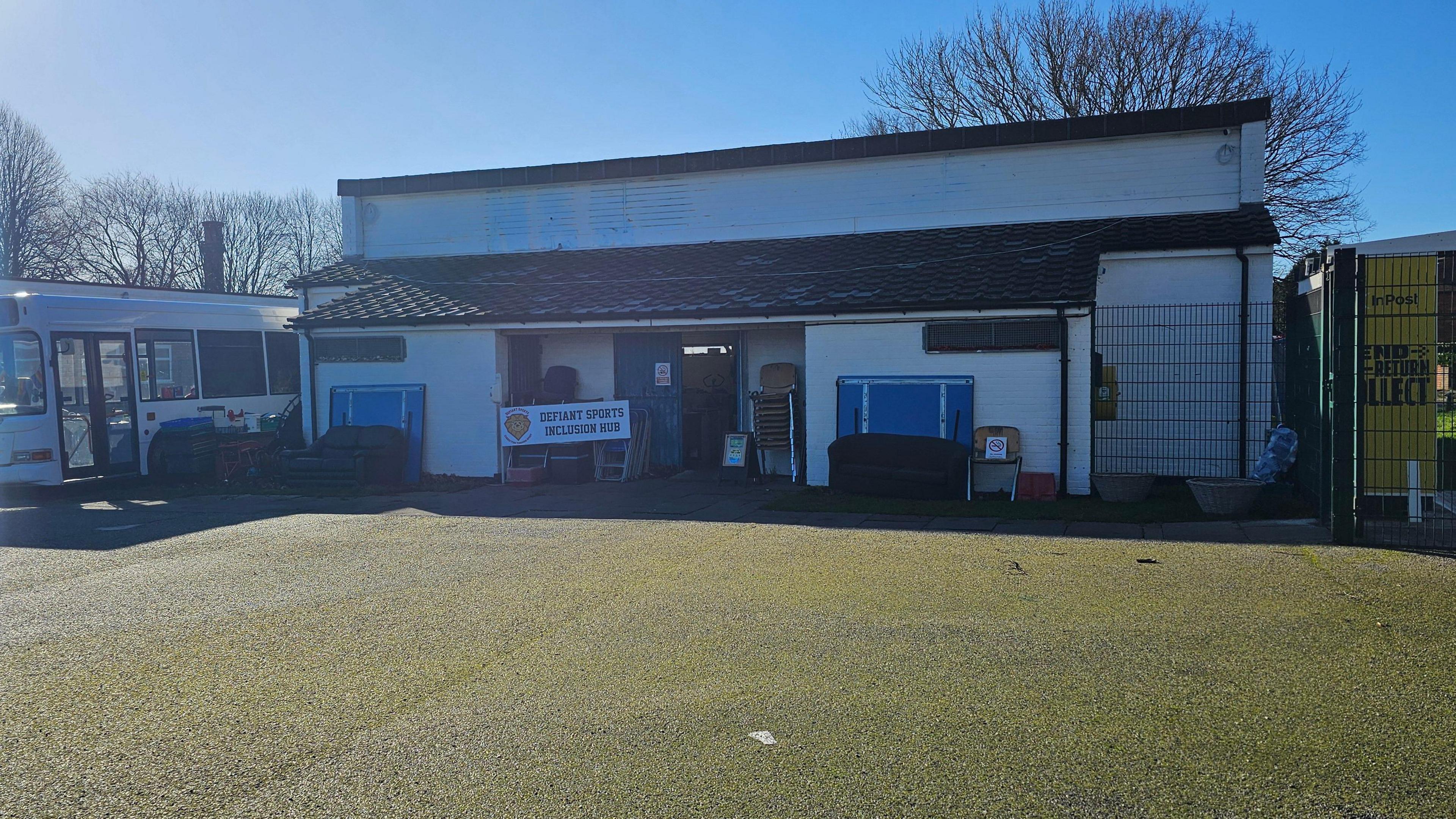 A white building with a bus next to it. They are on green grass patch under clear blue skies. 