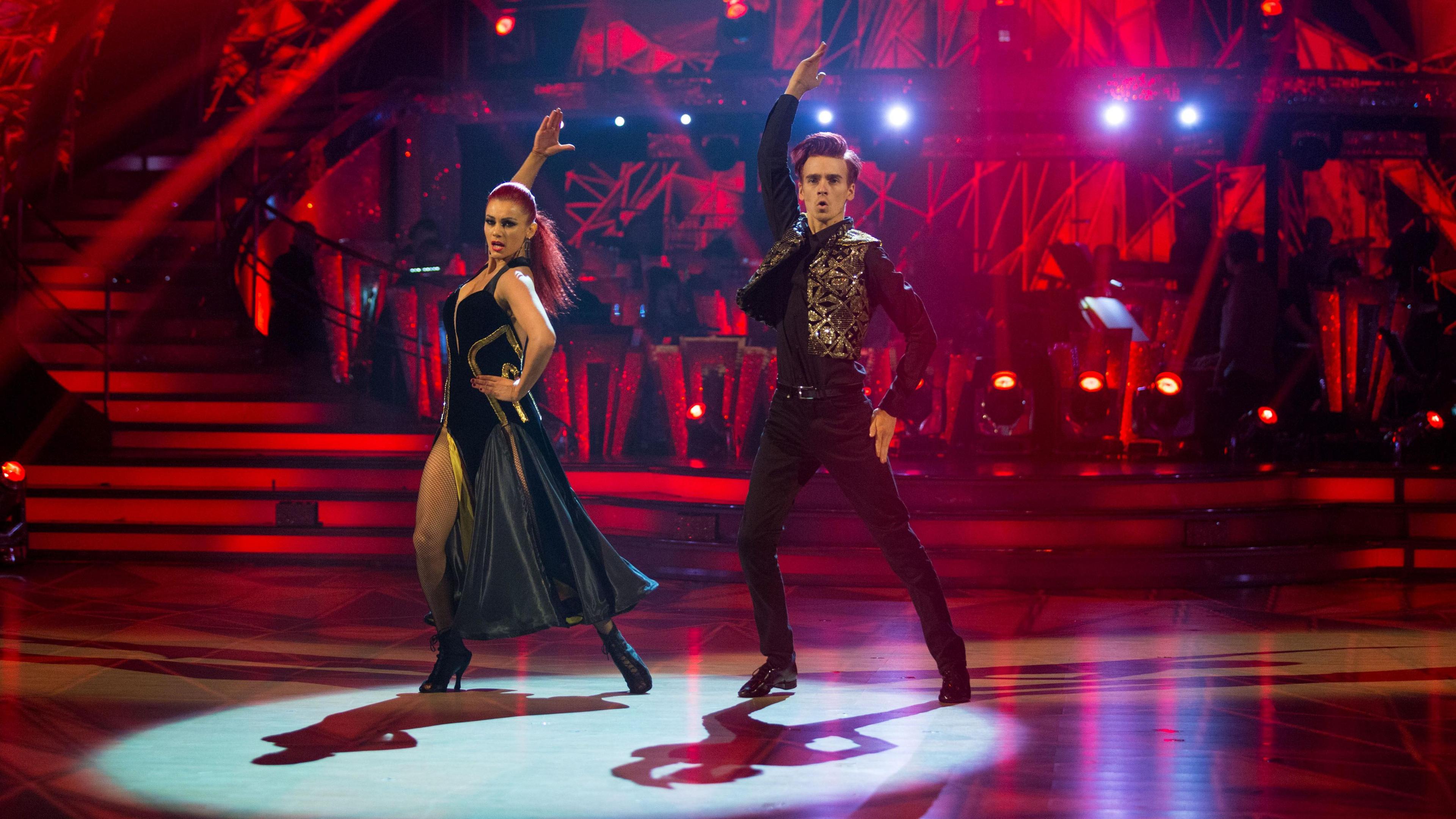 Dianne (L) and Joe (R) dance a Paso Doble. The stage lighting is red and there is a spotlight on them as they throw one arm up in the air