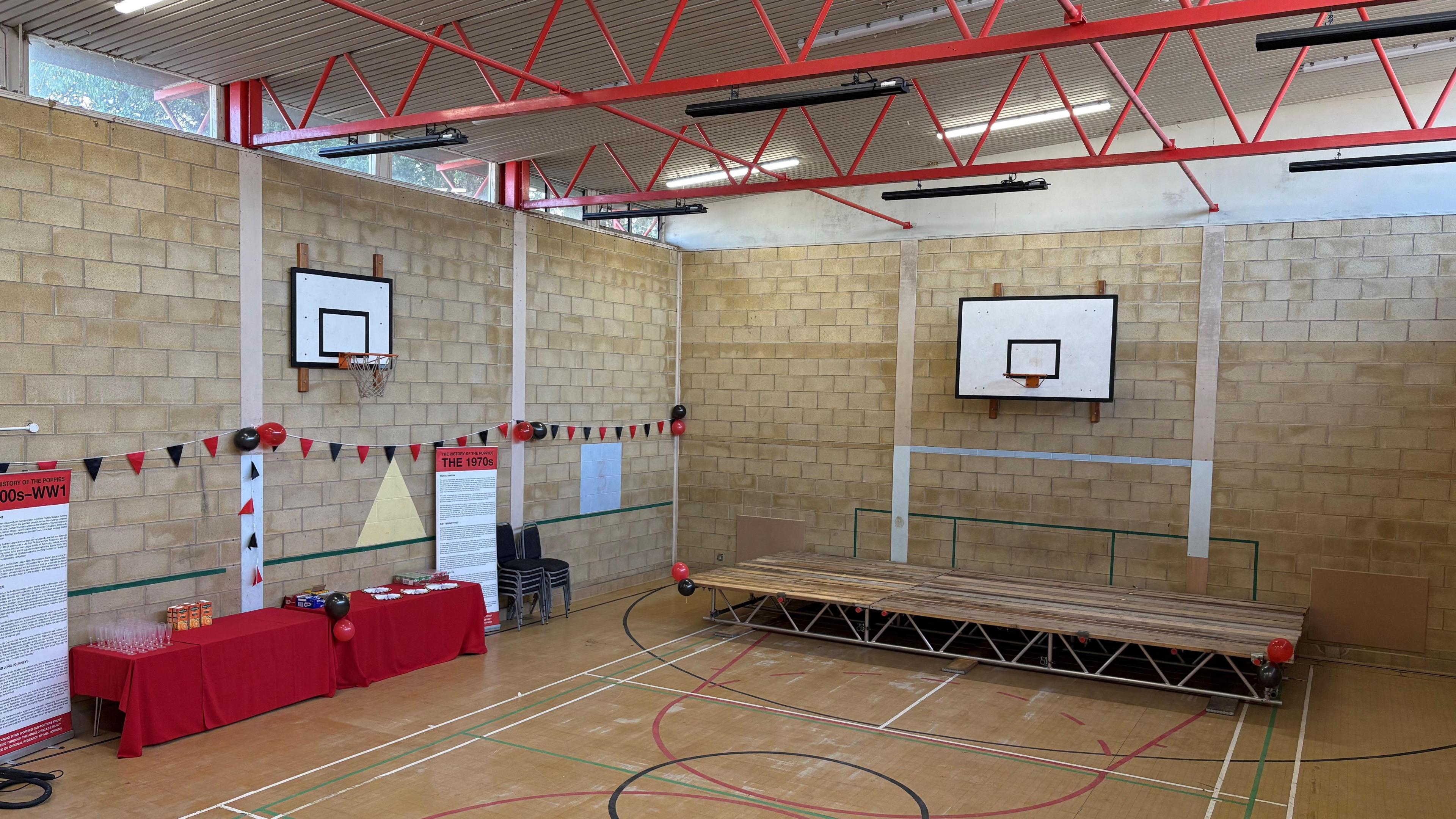 A community centre, that looks like a sports hall, complete with a temporary stage, two tables with red clothes on them, some bunting up, some signs, two basket ball hoops on the wall and red metal frames in the roof.