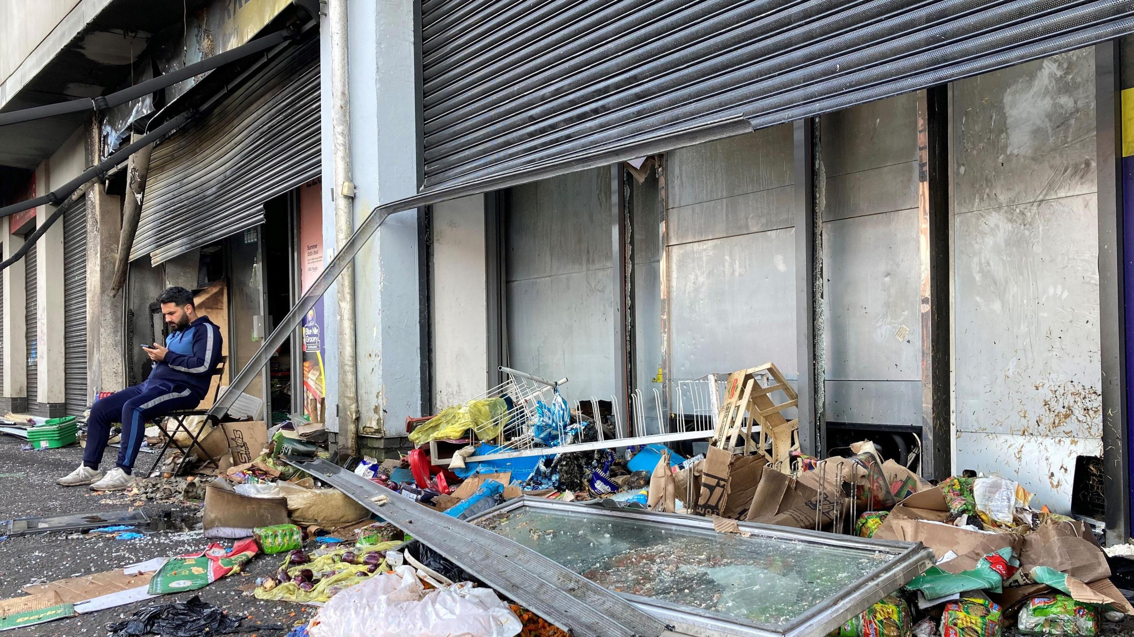 Front of burnt out business and the owner sitting on a chair in the street