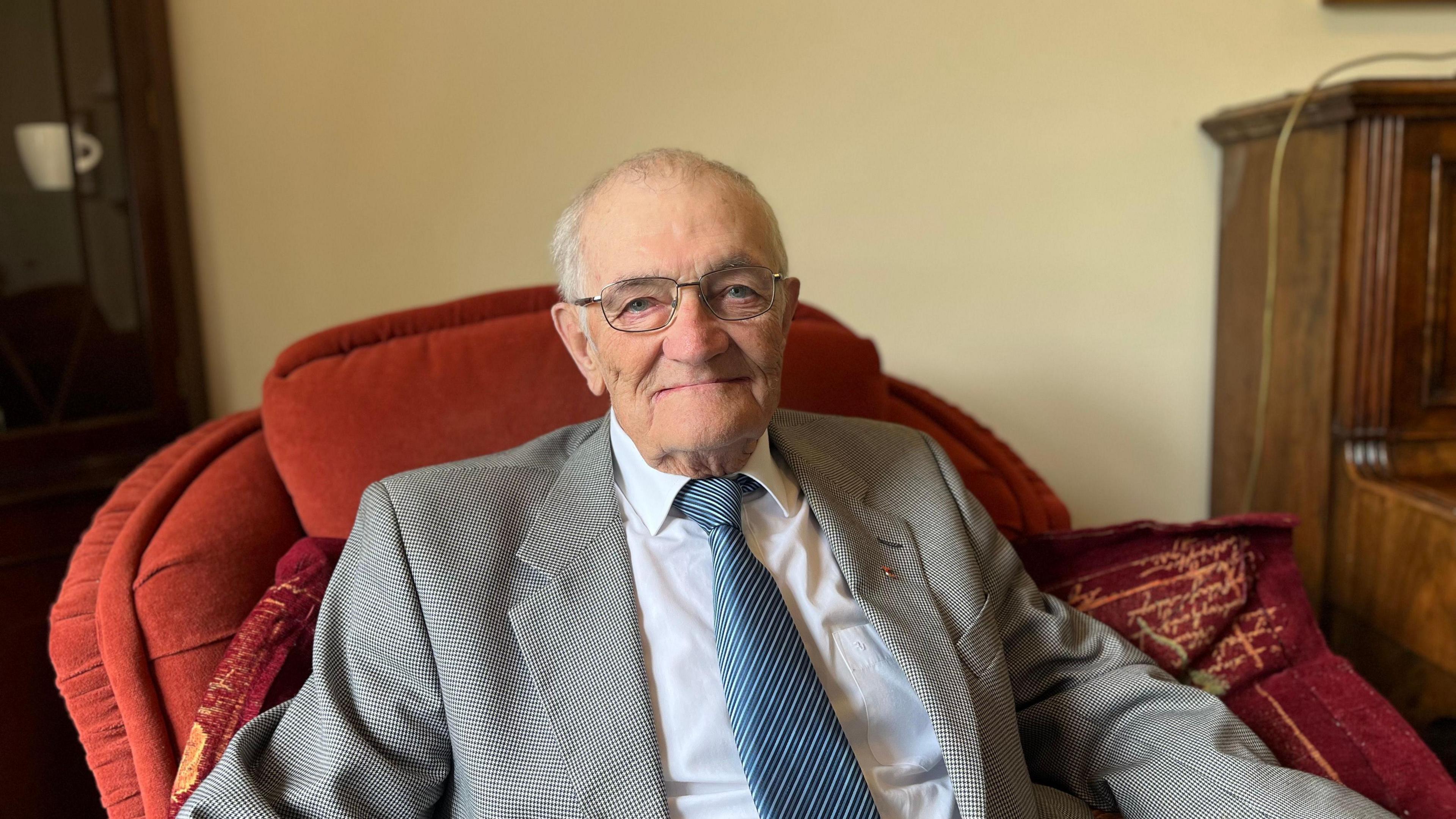 a man with grey hair and glasses sitting in a comfortable looking armchair and smiling into the camera