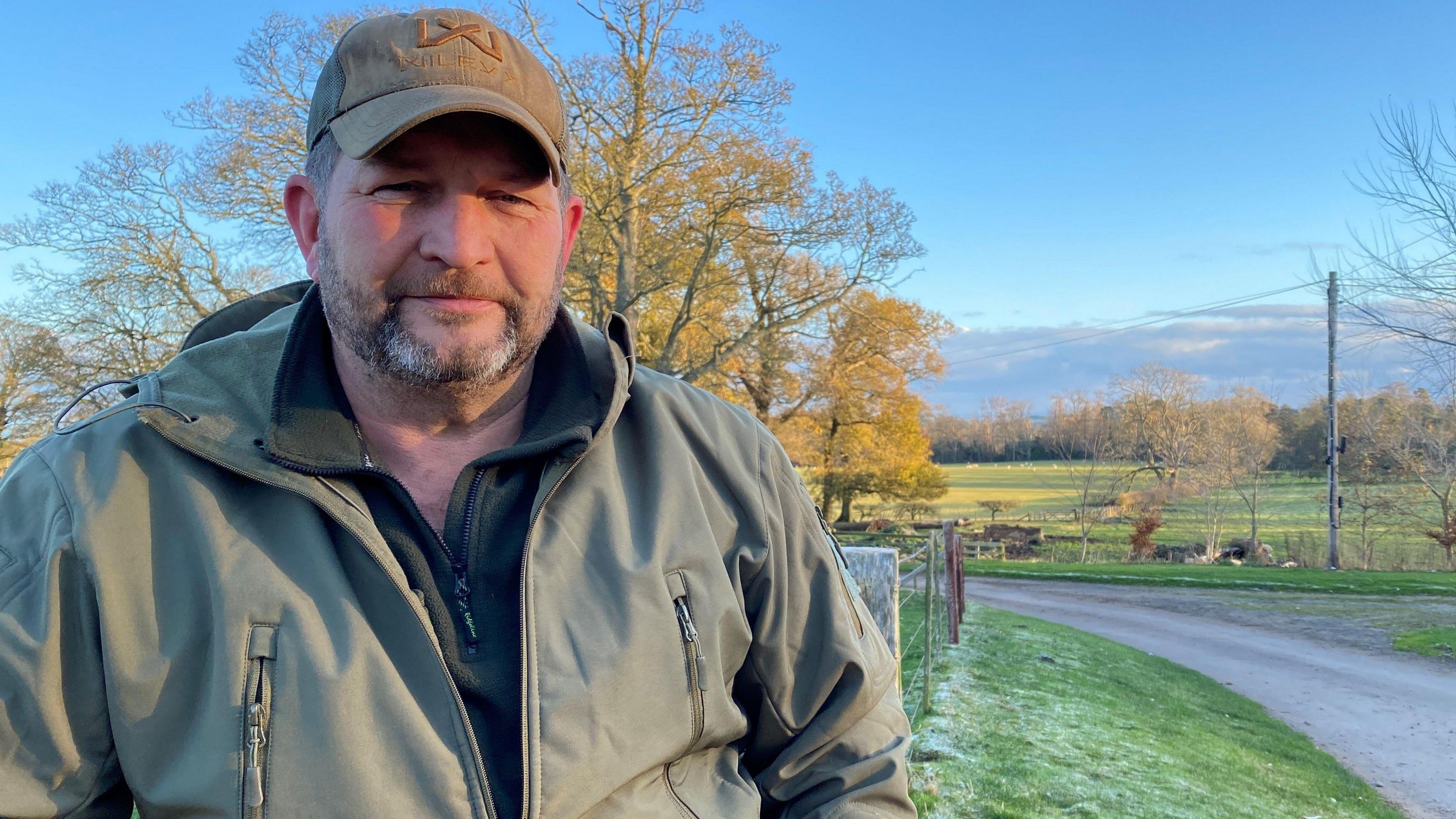 Daniel Spours is standing by a gate with fields of sheep behind him. He has a short stubbly beard and wear a cap.