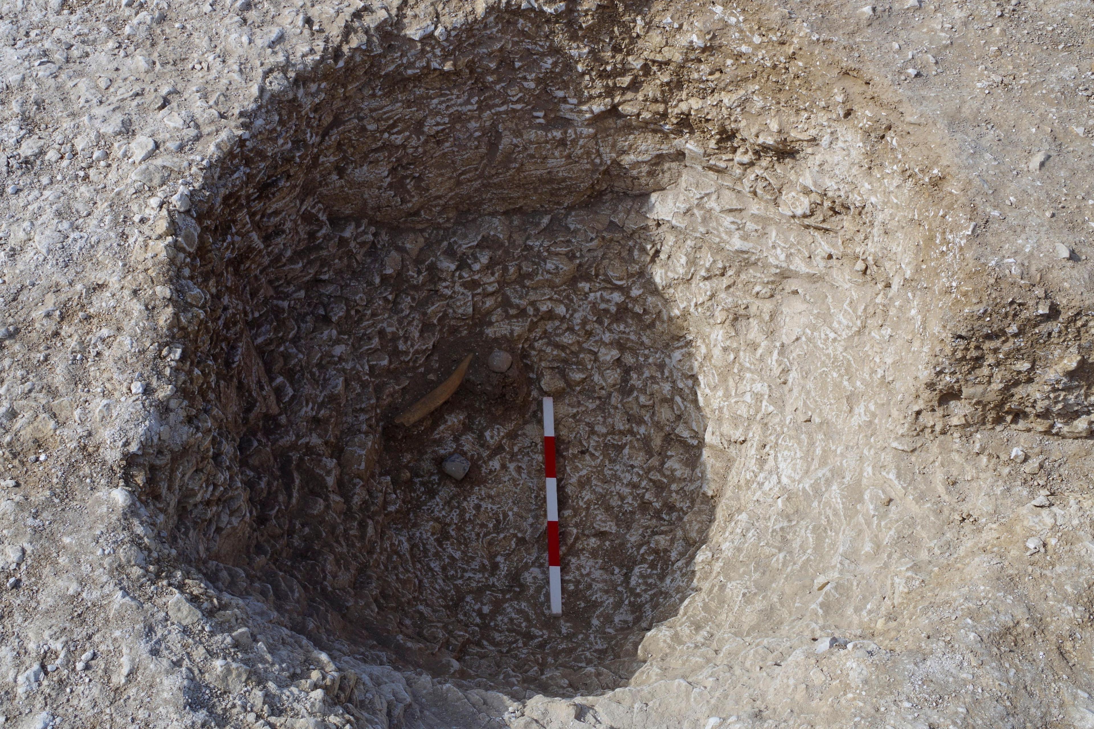 The hole in the ground shows a light rock with a hole in shadow dug into it. At the bottom is a red and white measuring pole, with three blocks of white and two blocks of red visible in between. It's difficult to see how deep it is but at the bottom there is an artefact protruding at the bottom on the left hand side of the hole. It is in the shadowed area and is something that looks light brown in colour and slightly curved. 