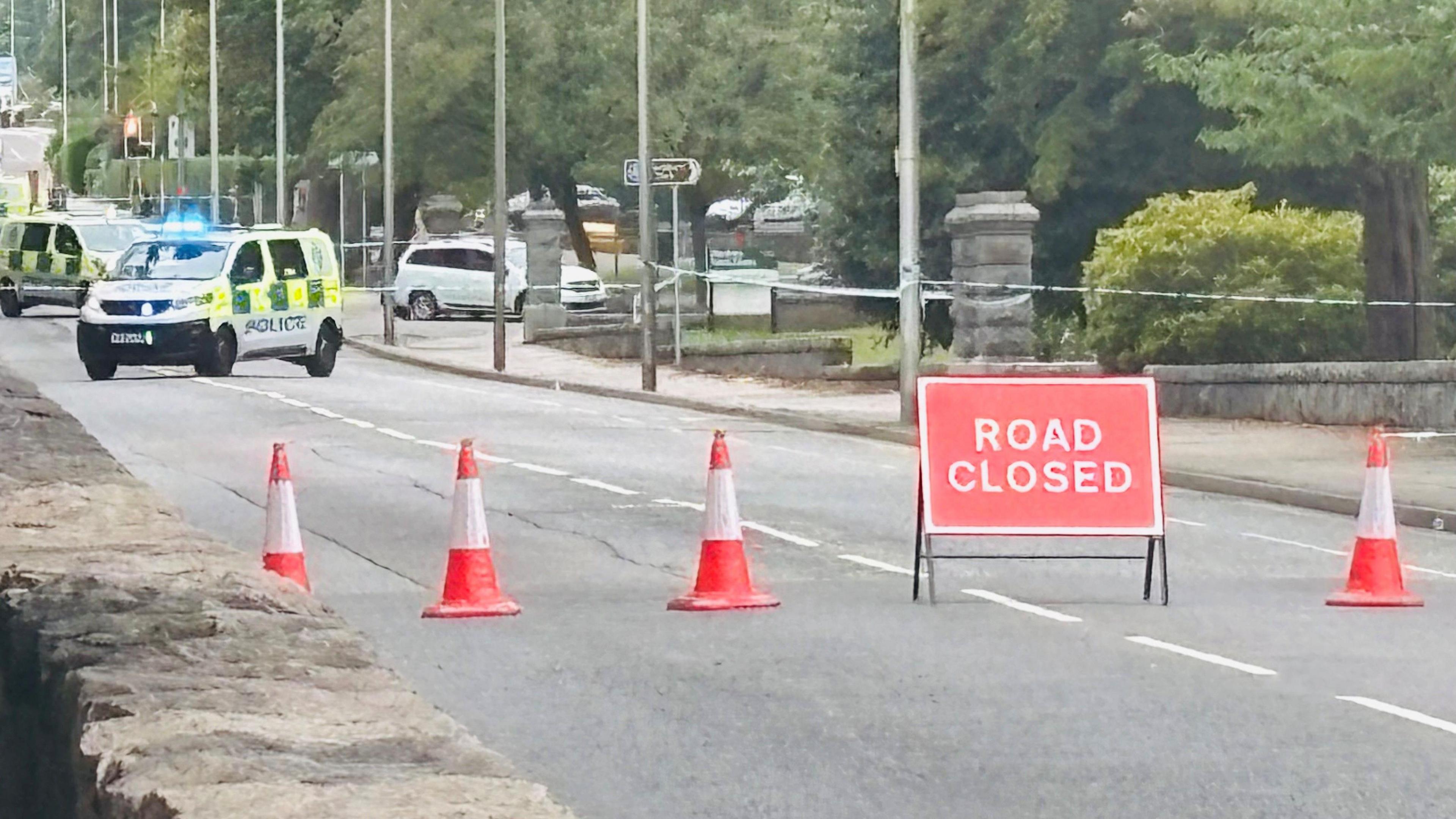 Police vehicles, police tape and police officers at scene of crash bike on road, in between two parks, with a road closed sign.